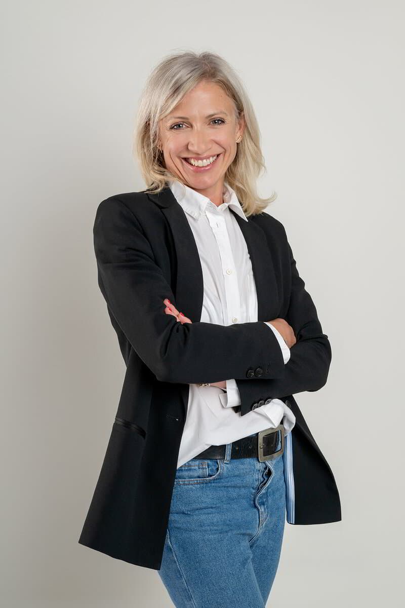 Une femme souriante en blazer noir, bras croisés, fond gris