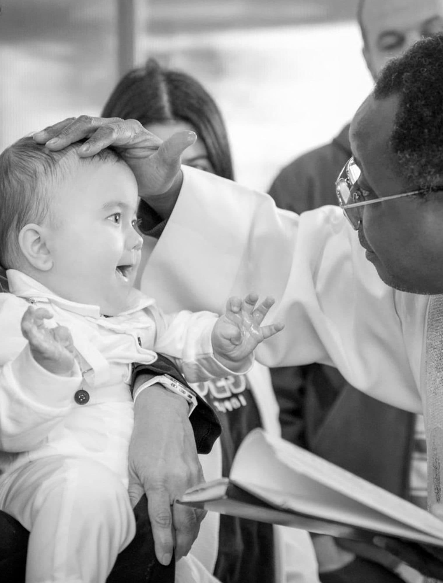 Un bébé éclate de rire sous la main du prêtre posée sur sa tête, en noir et blanc