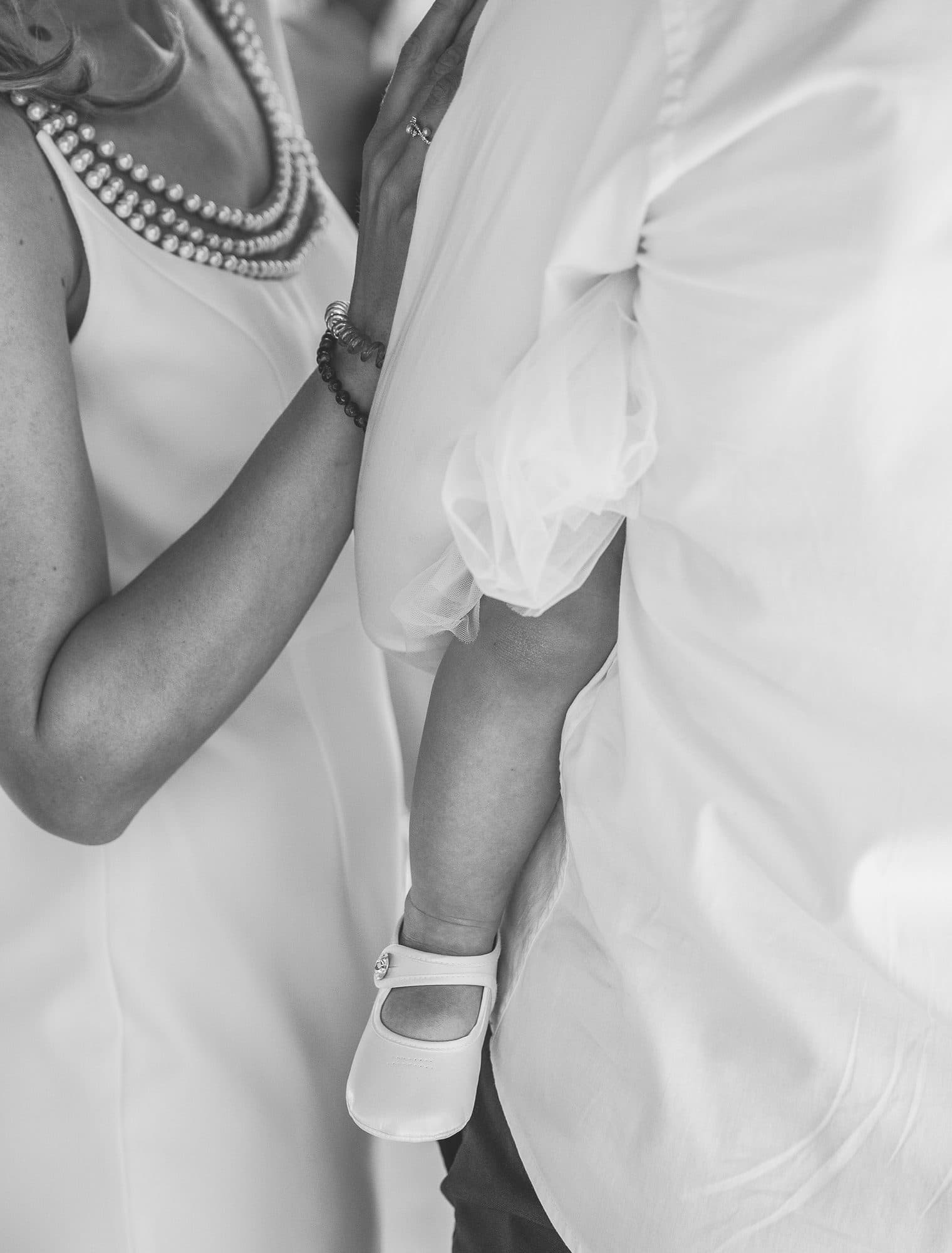 Le bras d’un bébé et sa petite chaussure blanche, contre sa maman au collier de perles, en noir et blanc
