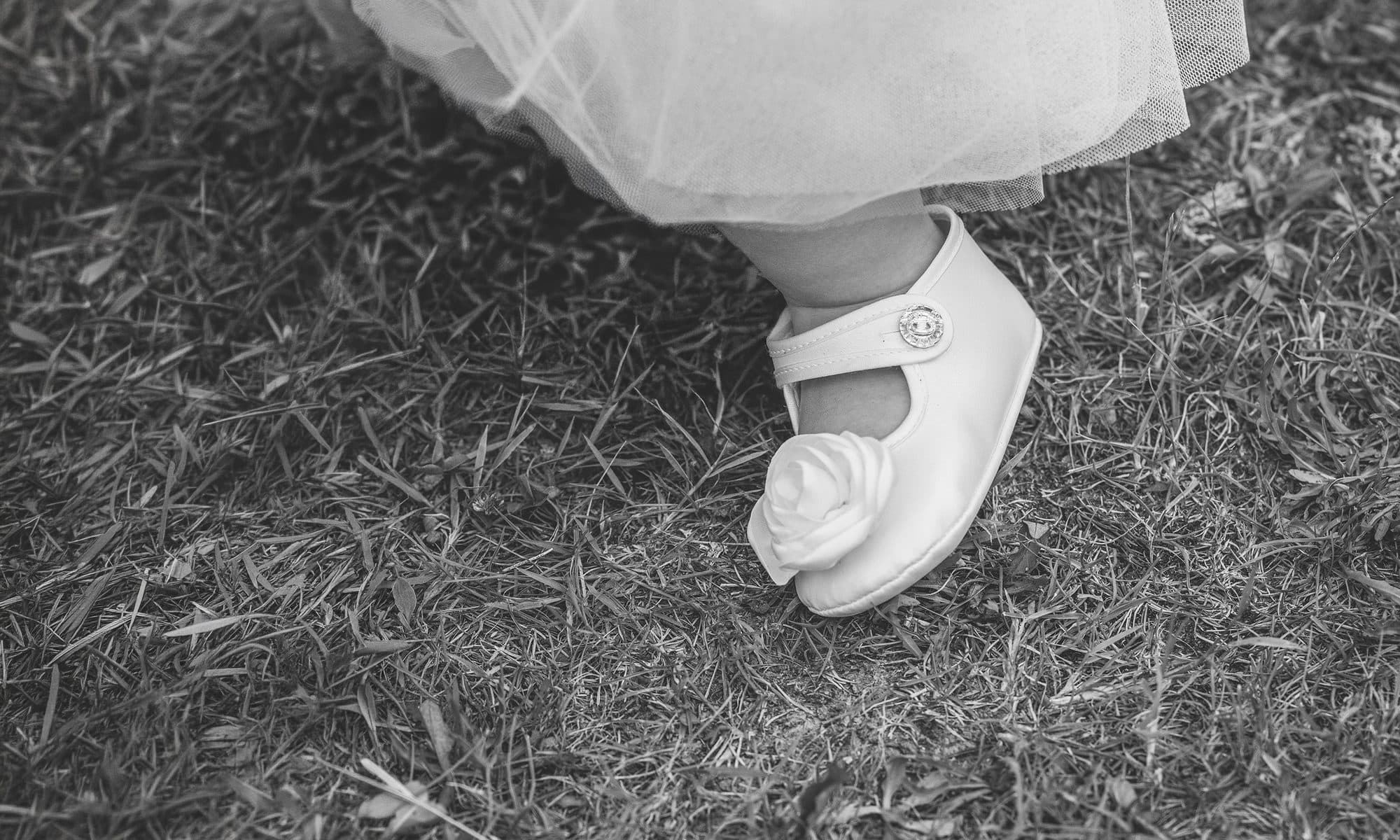 La chaussure blanche d’une petite fille sous sa robe de tulle, posée sur l’herbe, en noir et blanc