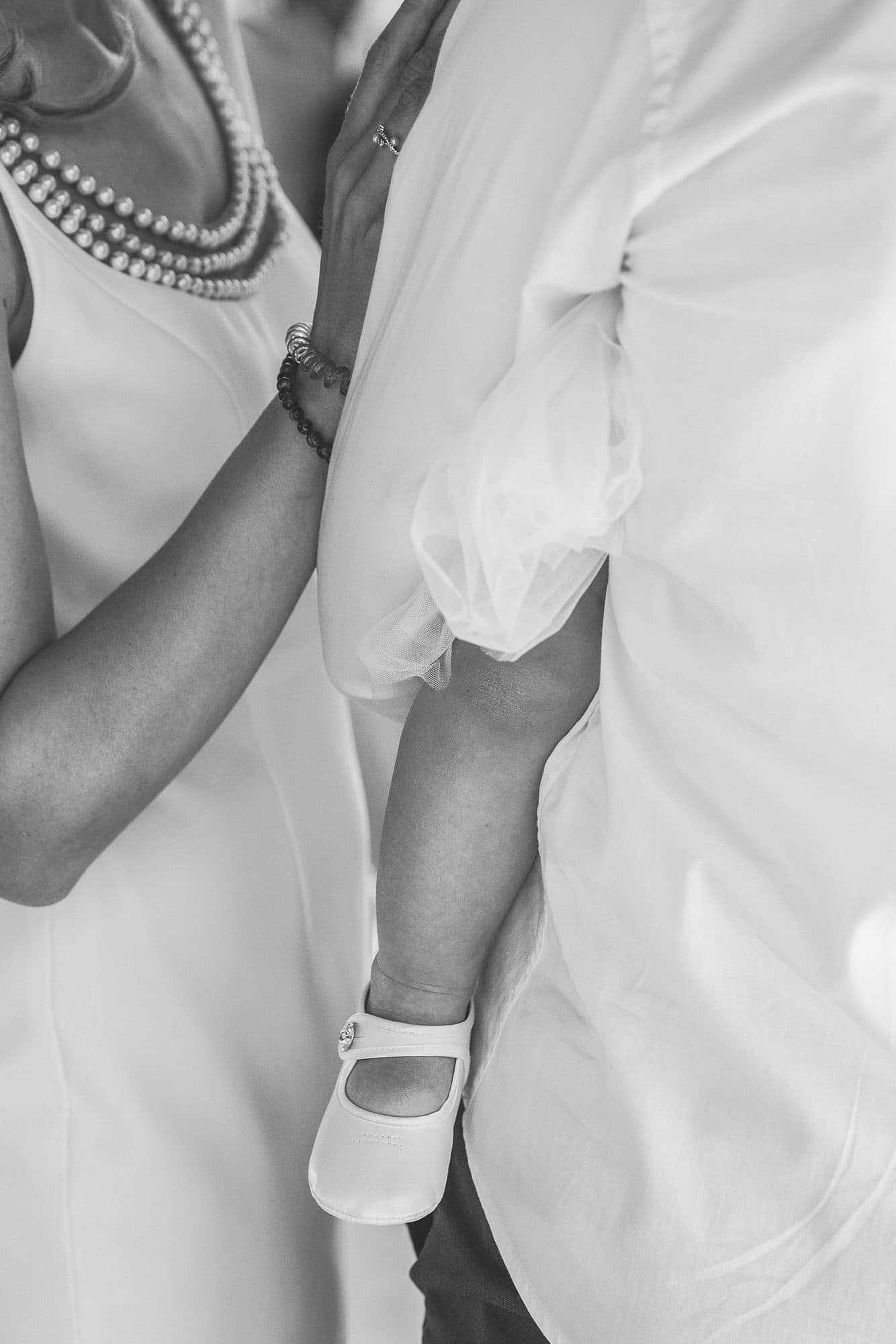 Le bras d’un bébé et sa petite chaussure blanche, contre sa maman au collier de perles, en noir et blanc