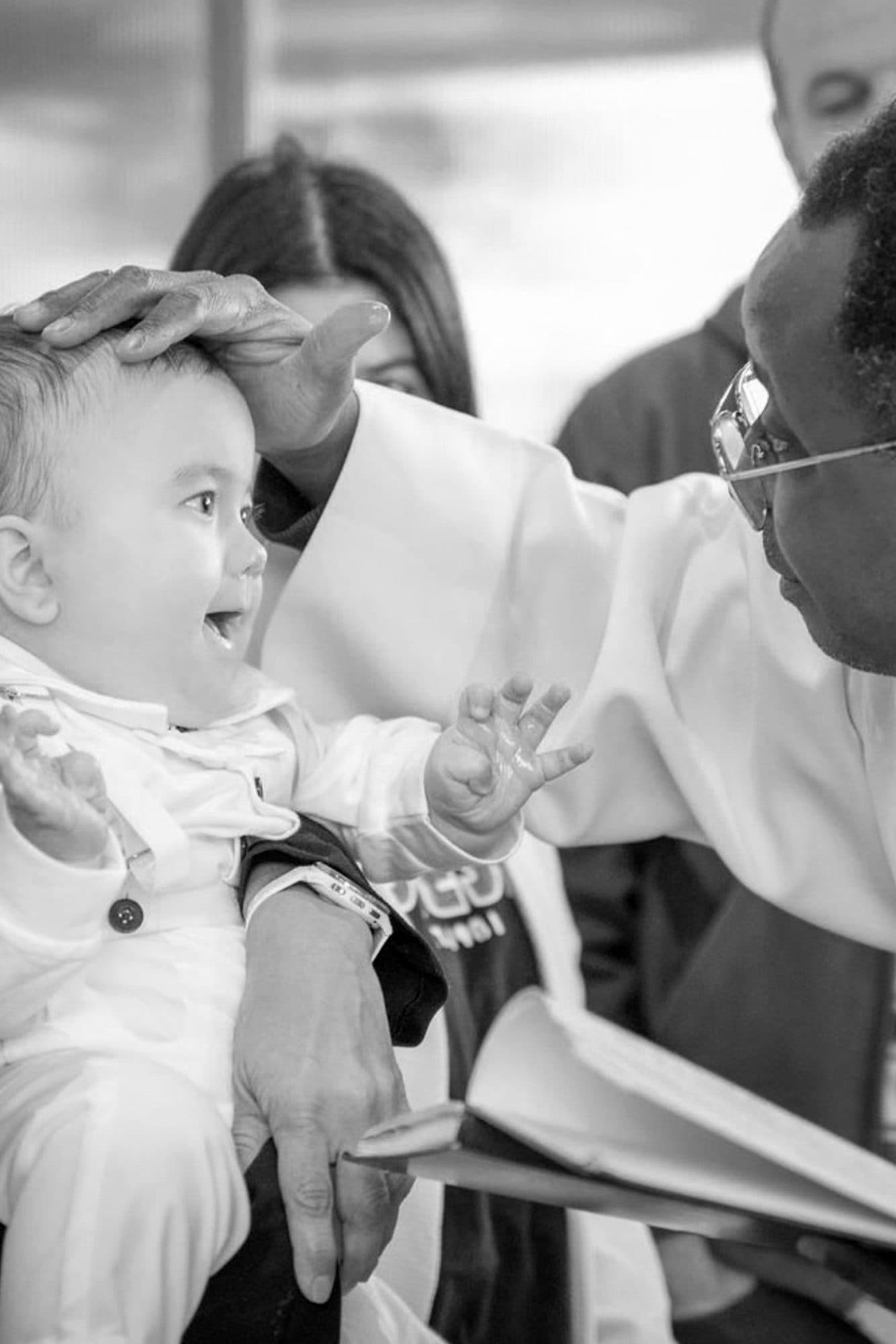 Un bébé éclate de rire sous la main du prêtre posée sur sa tête, en noir et blanc