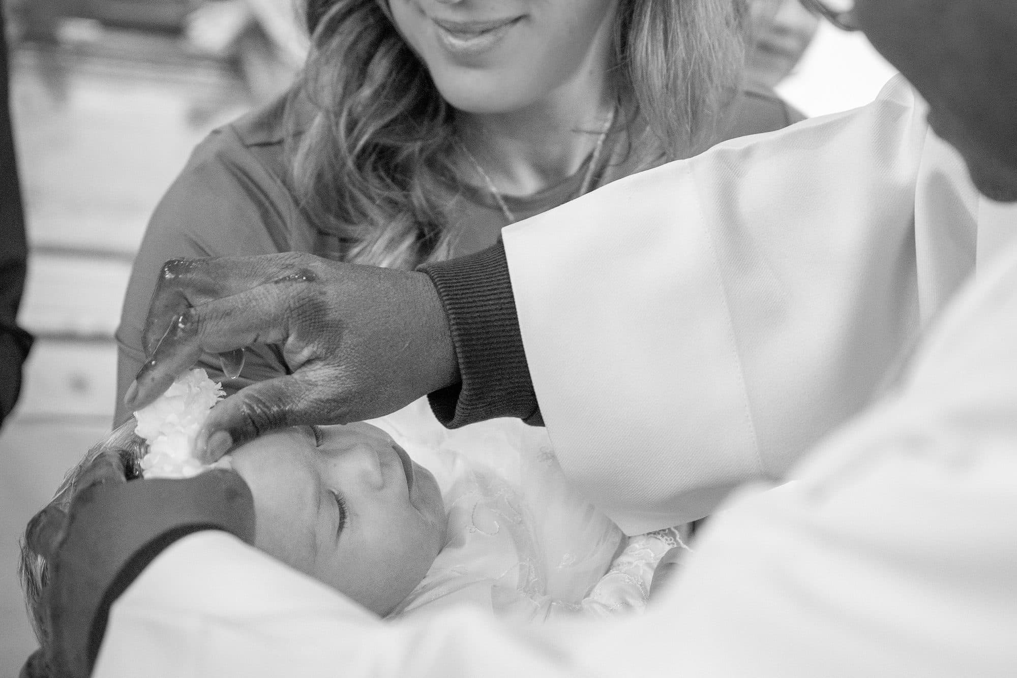 Le prêtre bénit le front d’un bébé endormi, sa maman souriante derrière lui, en noir et blanc
