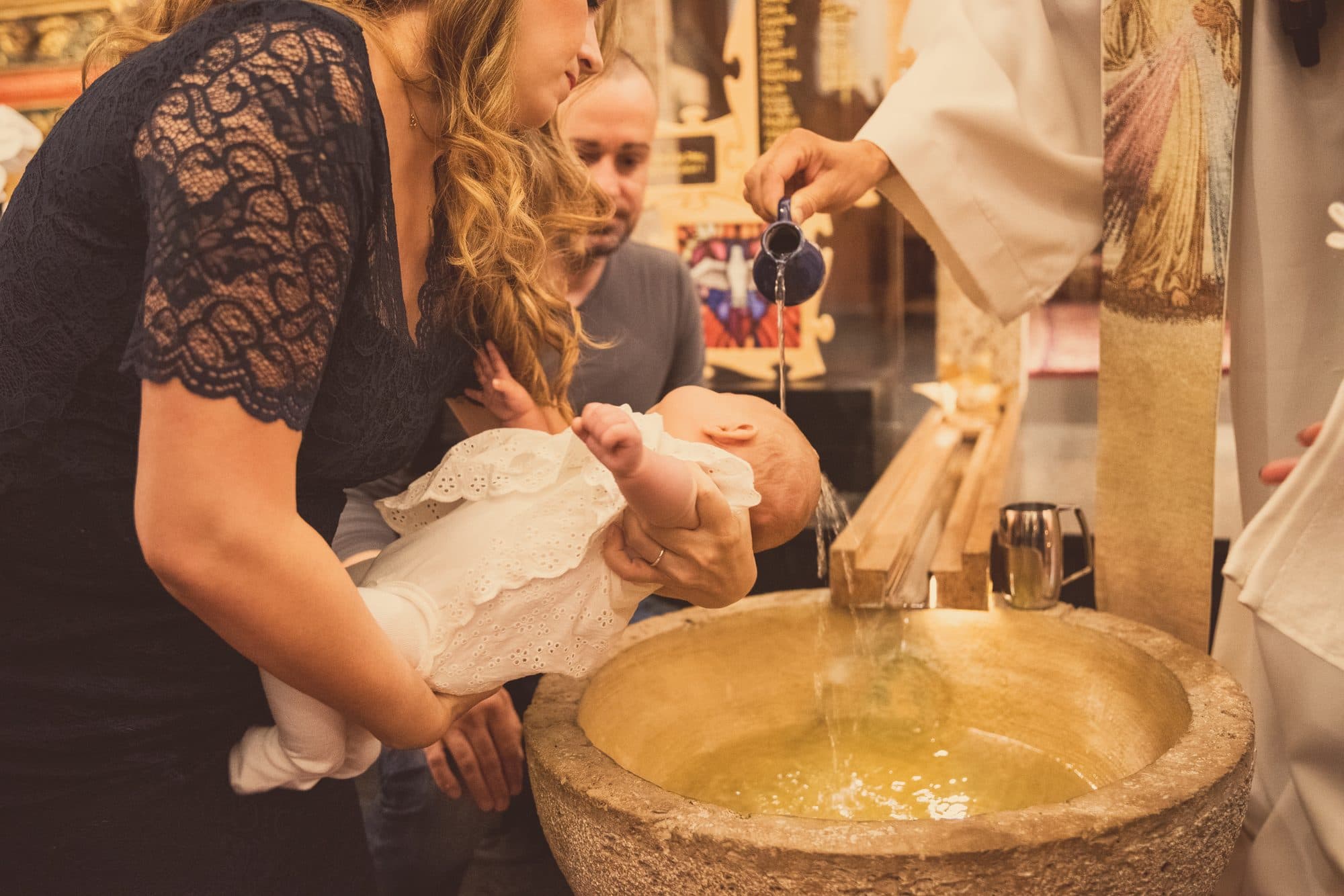 La maman tient son bébé au-dessus des fonts baptismaux pendant que le prêtre verse l’eau