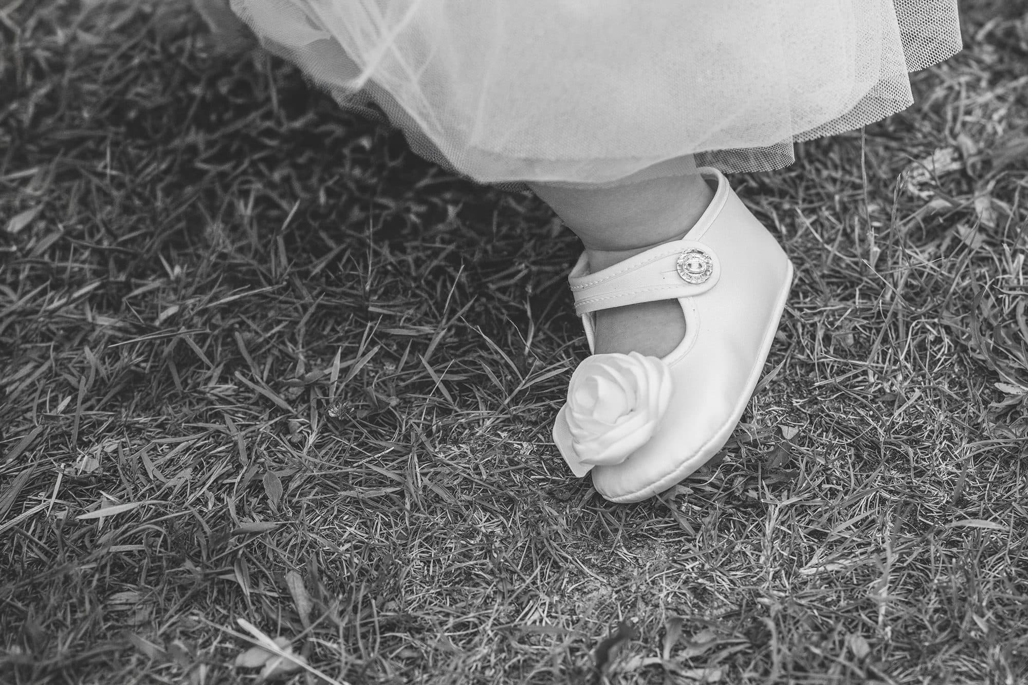 La chaussure blanche d’une petite fille sous sa robe de tulle, posée sur l’herbe, en noir et blanc