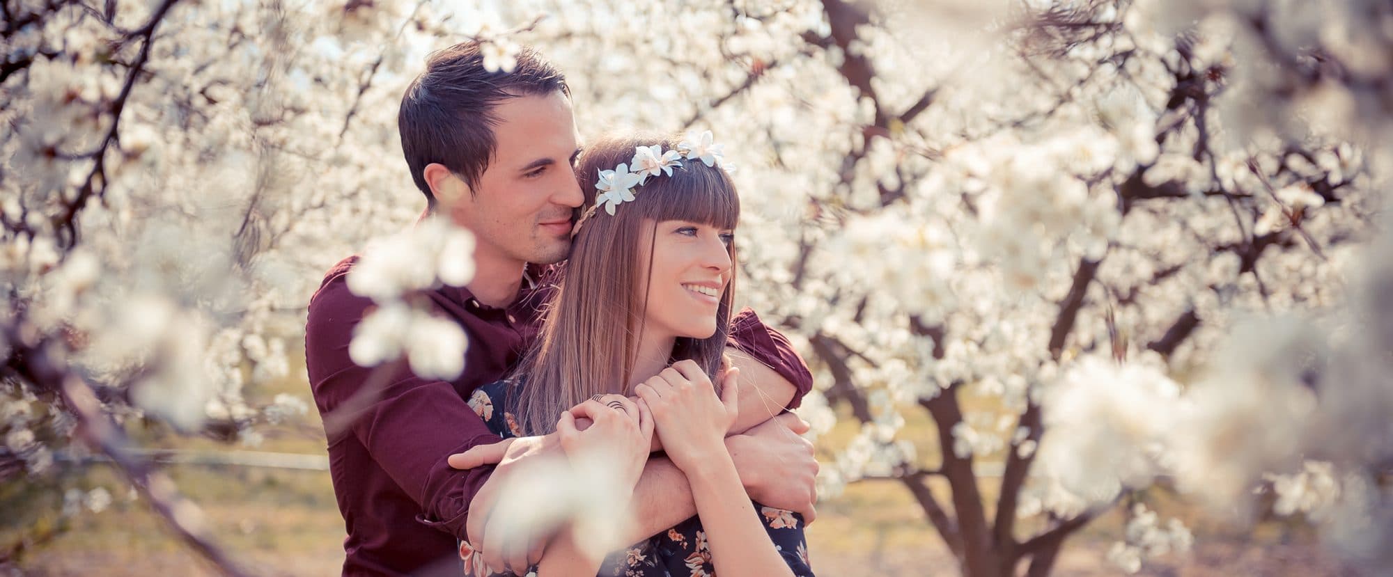 Un couple enlacé et souriant sous des cerisiers en fleurs, elle porte une couronne de fleurs blanches