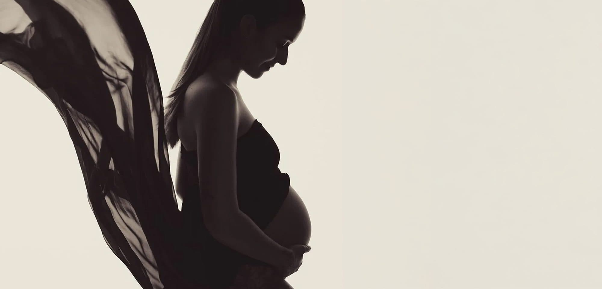La silhouette d’une future maman de profil, les mains sous le ventre, un voile sombre qui s’envole, sur fond crème
