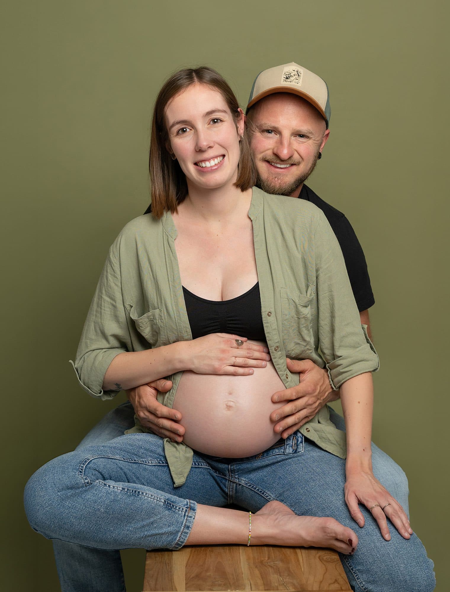 Une future maman assise sur un tabouret en bois, les mains sur le ventre, son conjoint en casquette derrière elle, sur fond vert olive