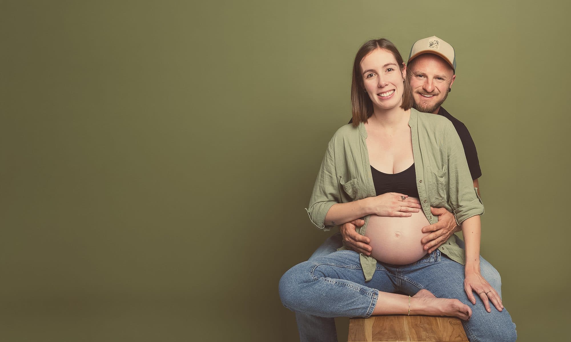 Une future maman assise sur un tabouret en bois, les mains sur le ventre, son conjoint en casquette derrière elle, sur fond vert olive