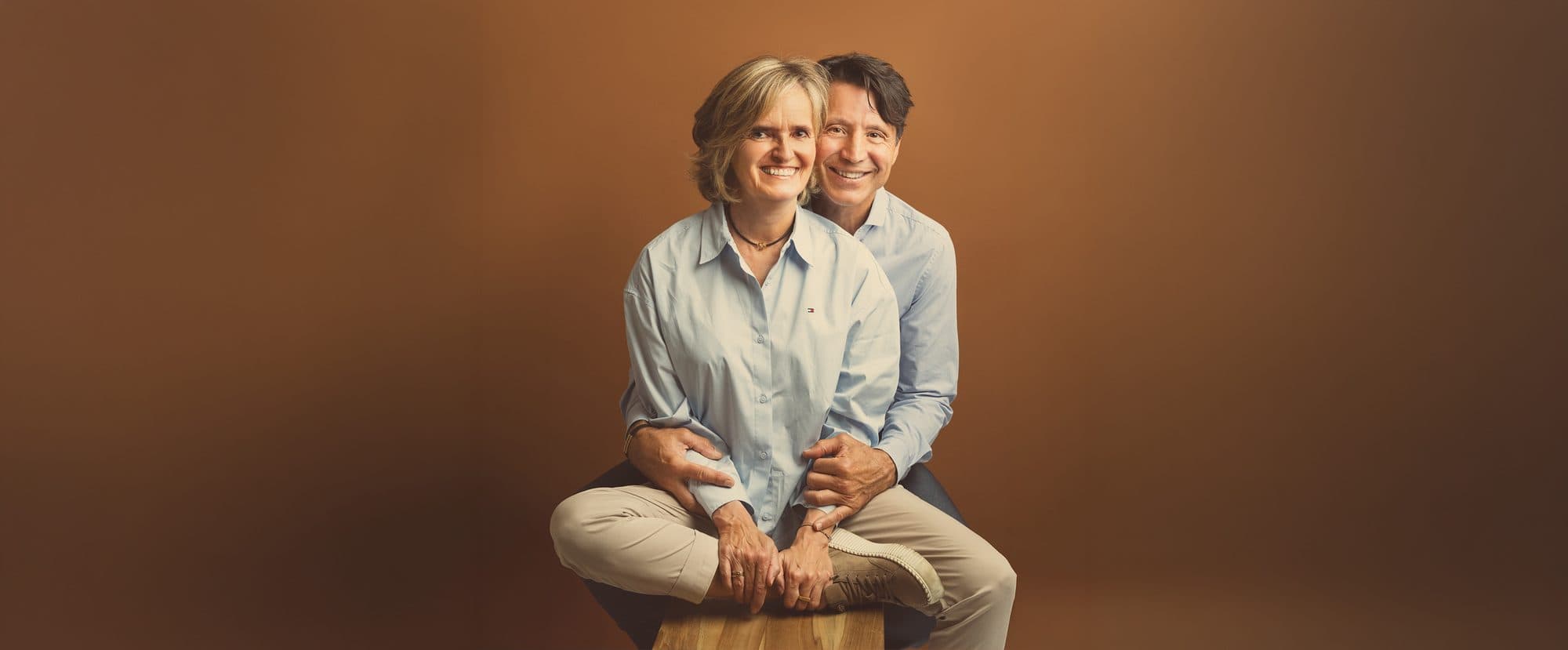 Un couple souriant assis l’un derrière l’autre sur un tabouret en bois, sur fond caramel