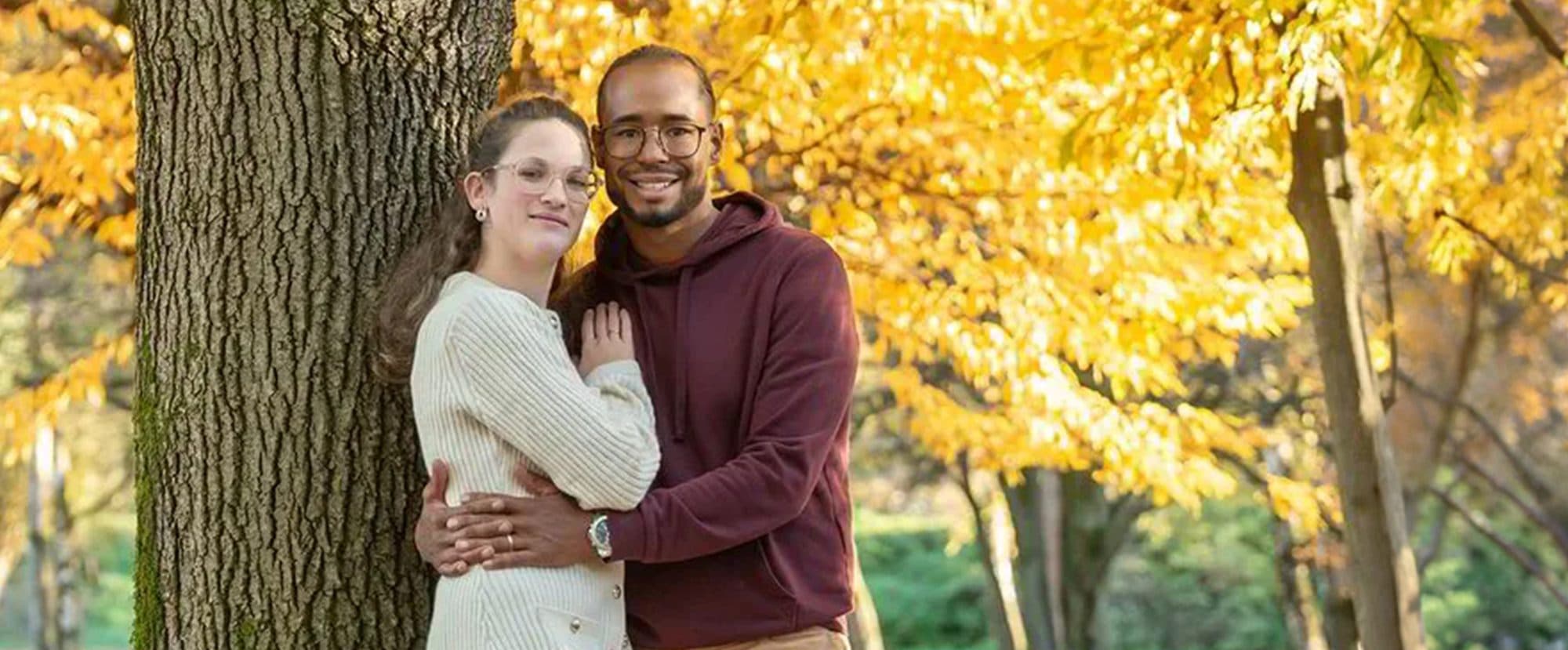 Un couple enlacé contre un grand tronc, sous un feuillage jaune d’automne