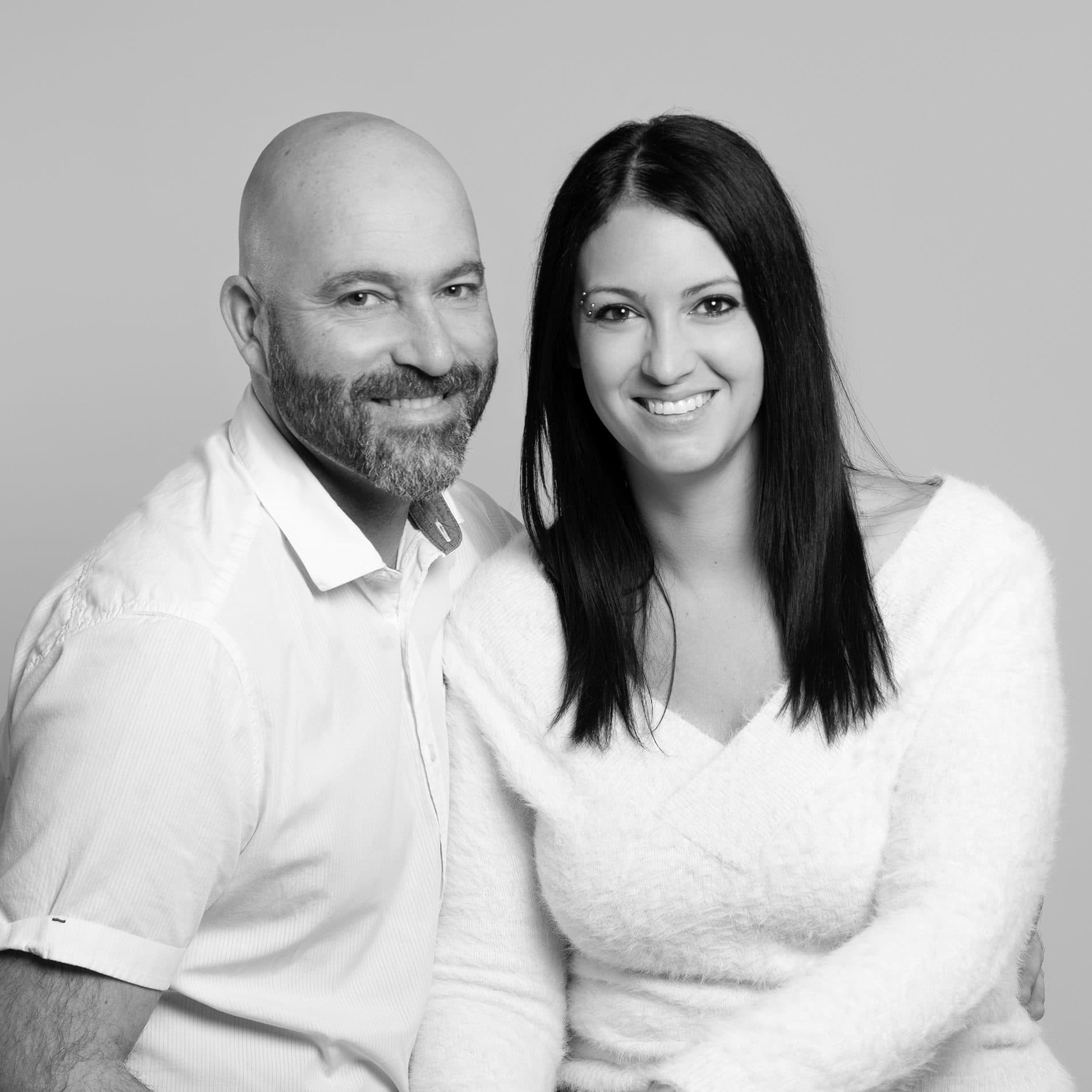 Un couple souriant habillé de blanc, côte à côte, en noir et blanc sur fond gris