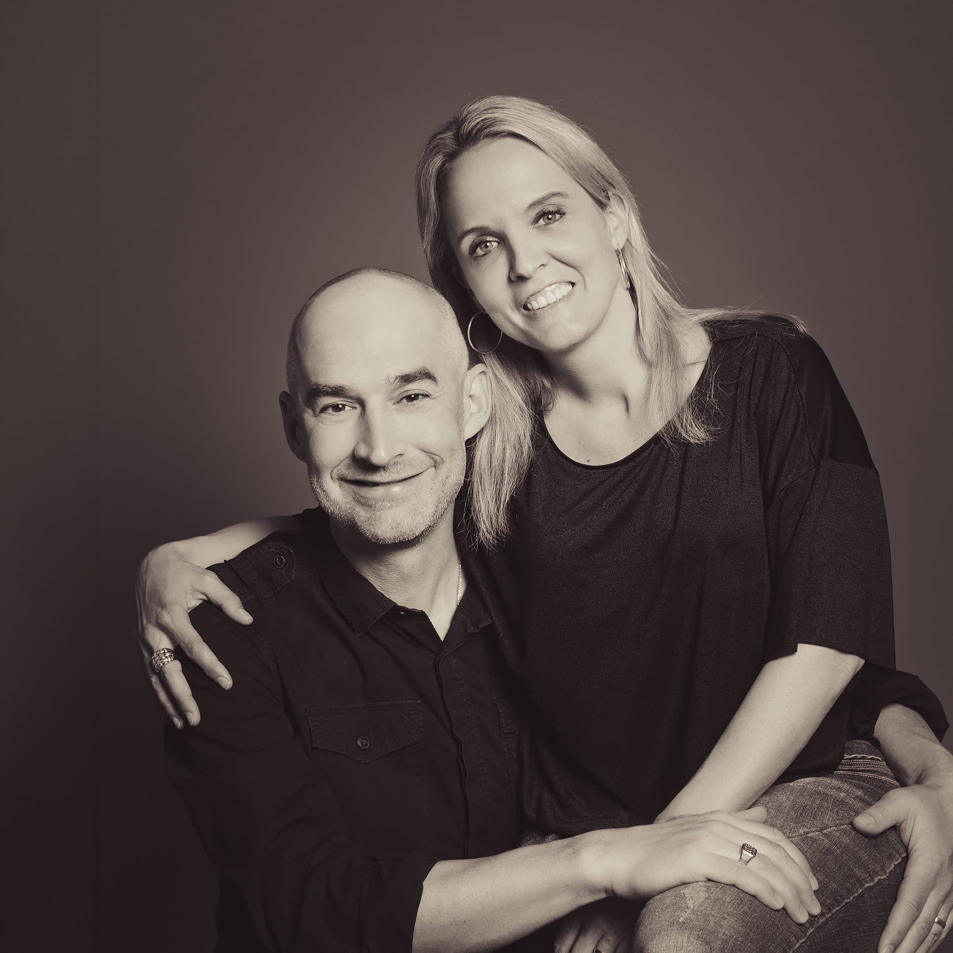 Un couple habillé de noir, assis l’un contre l’autre, en tons sépia sur fond sombre