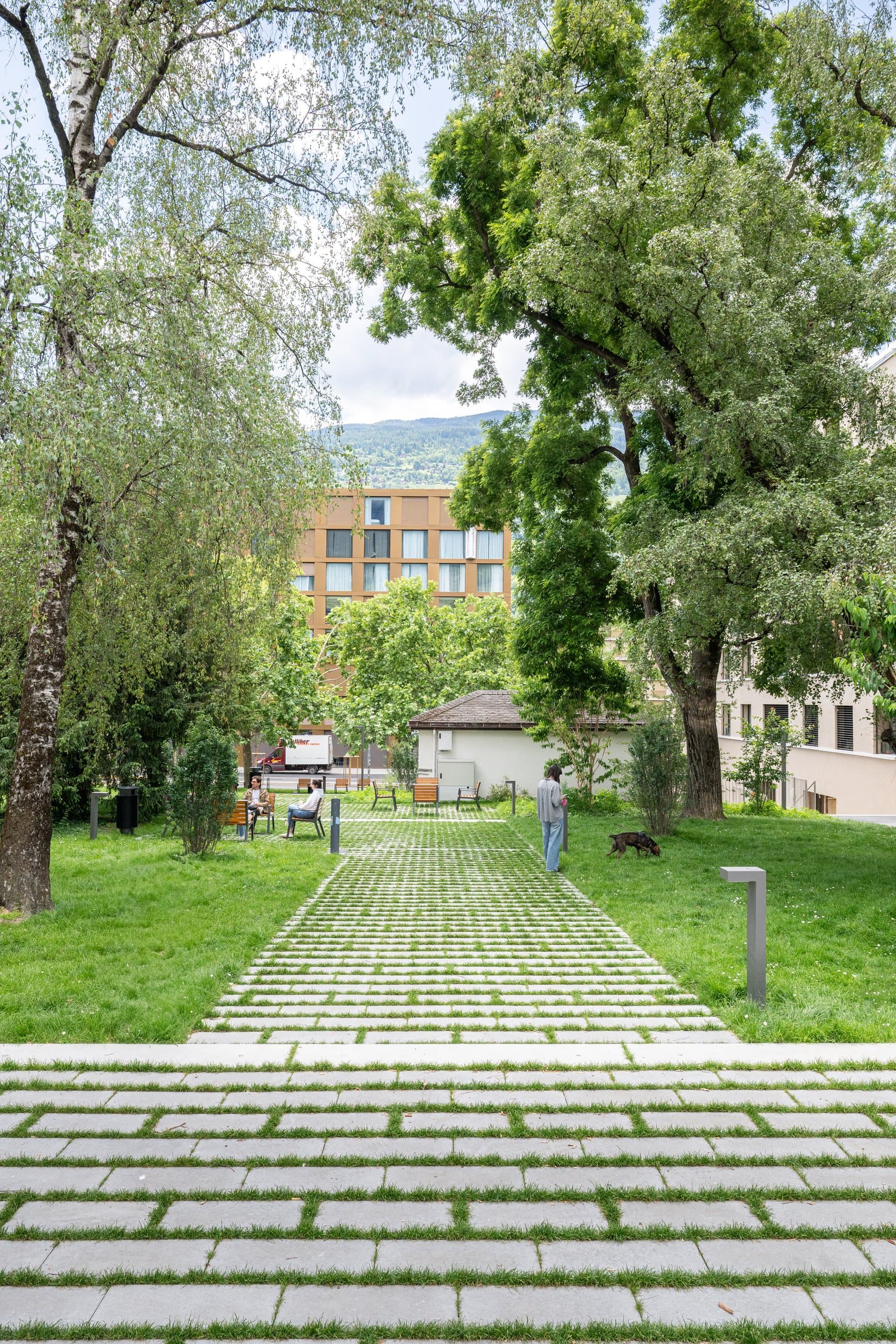 Allée de dalles engazonnées qui file entre deux pelouses et de grands arbres, au square des Mayennets à Sion