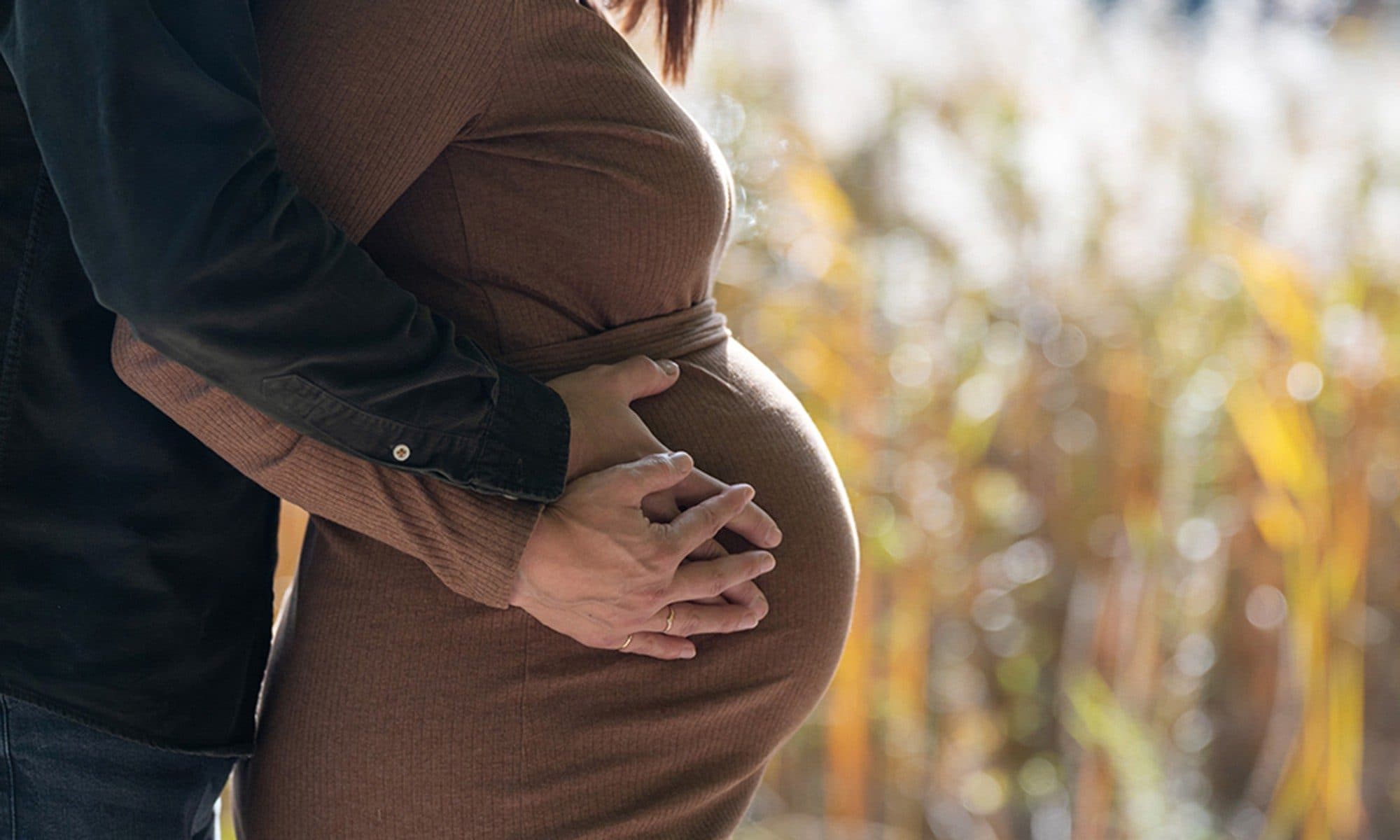 Un futur papa enlace sa compagne enceinte, leurs mains posées sur son ventre, devant des herbes dorées d’automne