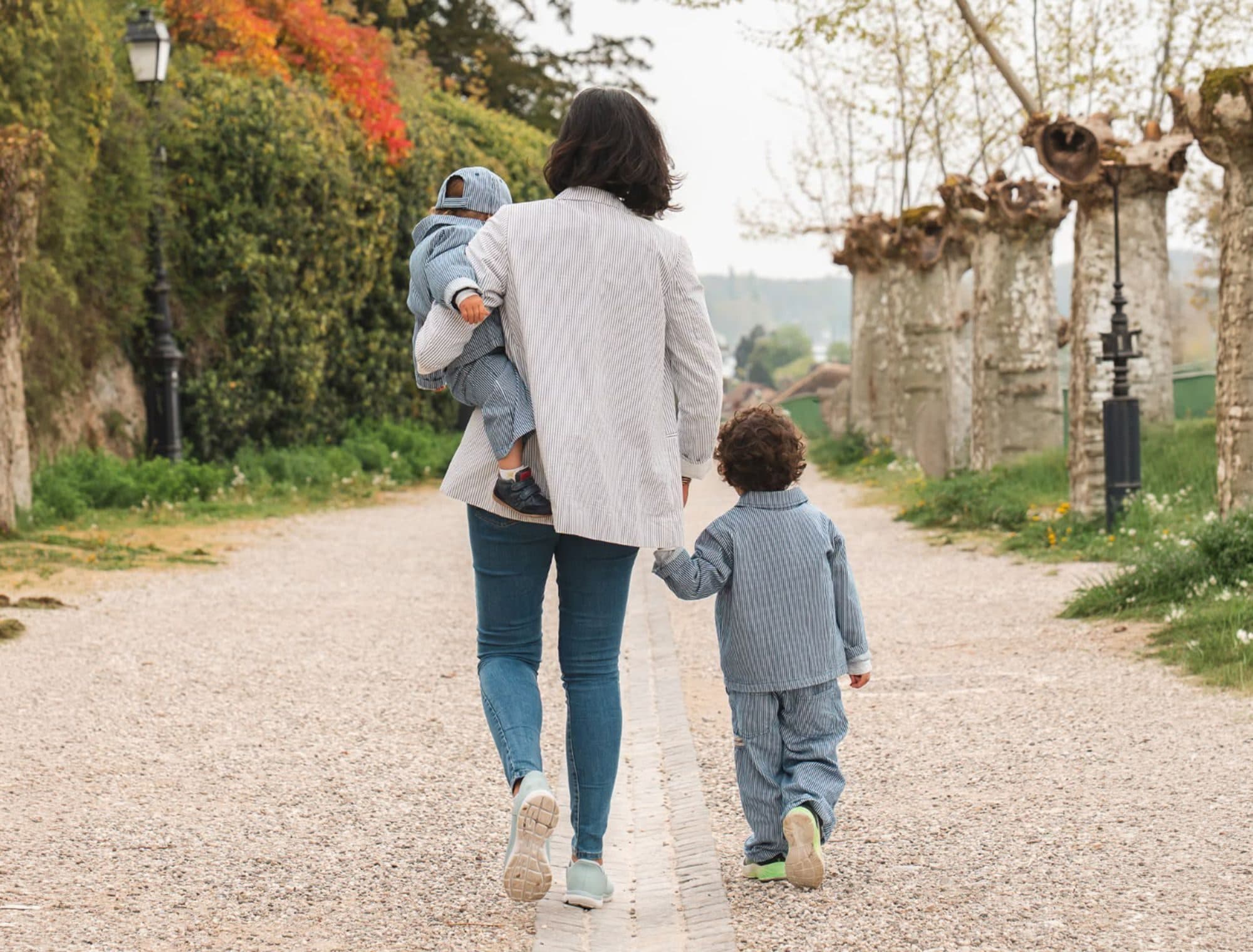 Une maman porte son bébé et tient son fils par la main, sur une allée bordée d’arbres taillés