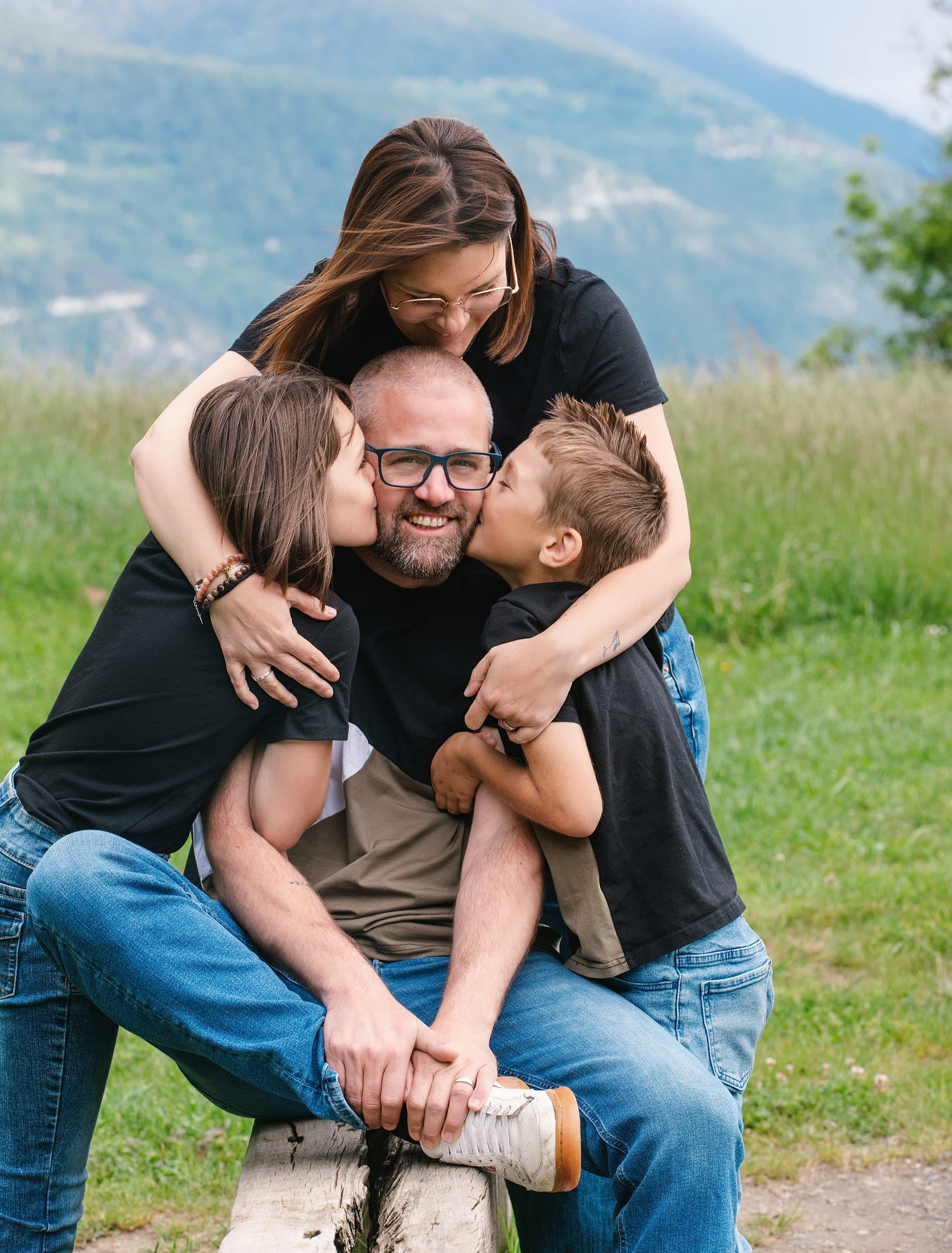 Un papa assis sur un banc dans l’herbe reçoit les bisous de sa fille et de son fils, la maman les enlace par derrière