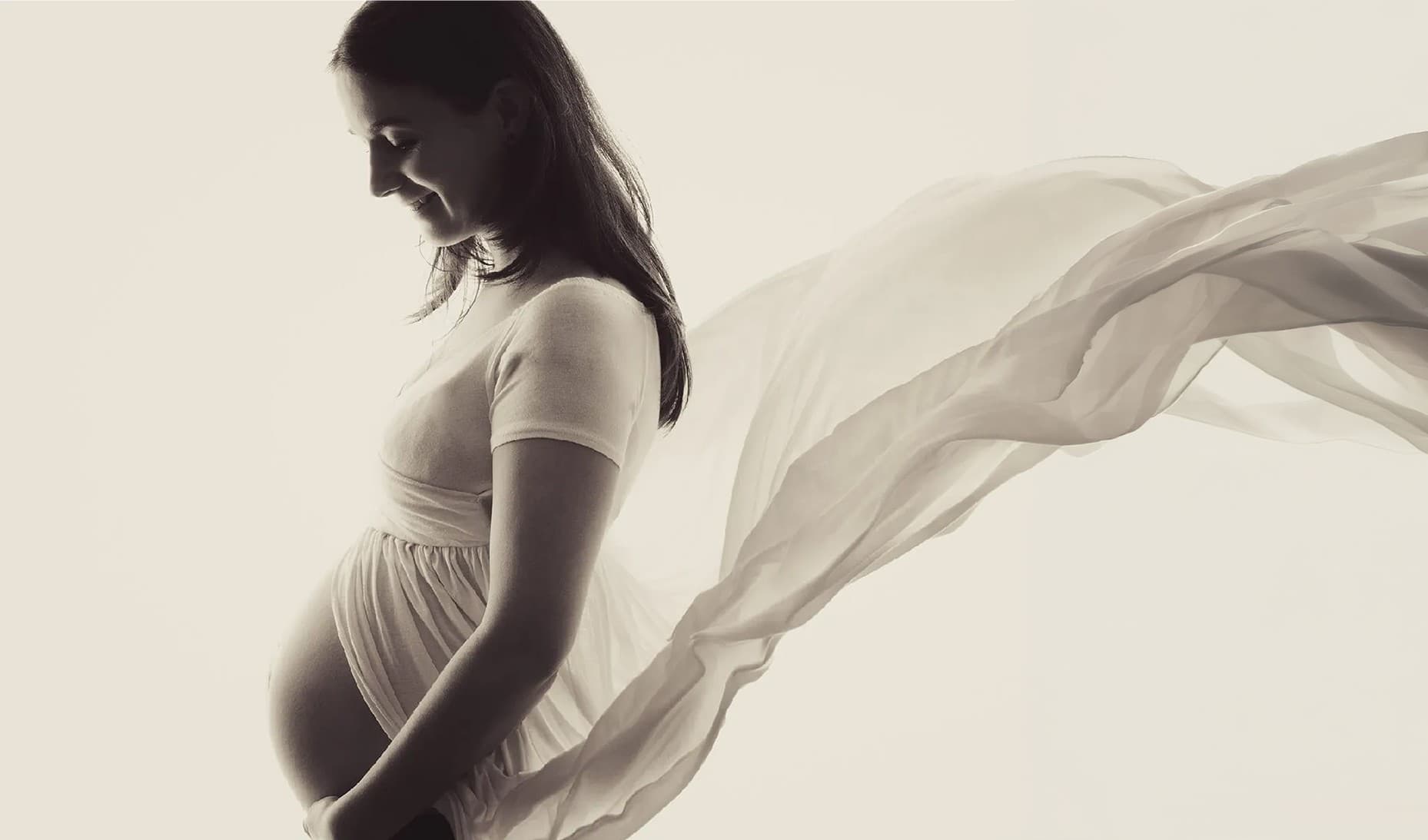 Future maman de profil en robe claire, un grand voile blanc soulevé derrière elle
