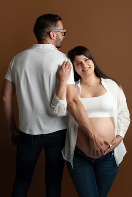 Future maman en jean et chemise blanche ouverte sur le ventre, son conjoint derrière elle, fond brun