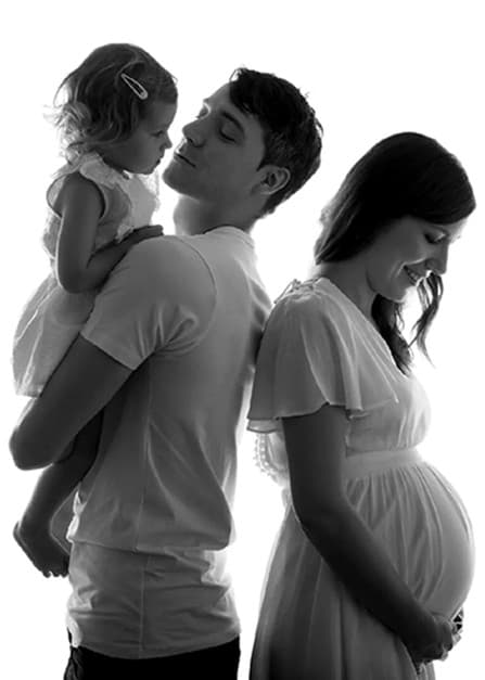 Future maman en robe claire, son conjoint en t-shirt porte leur fillette, en noir et blanc sur fond blanc