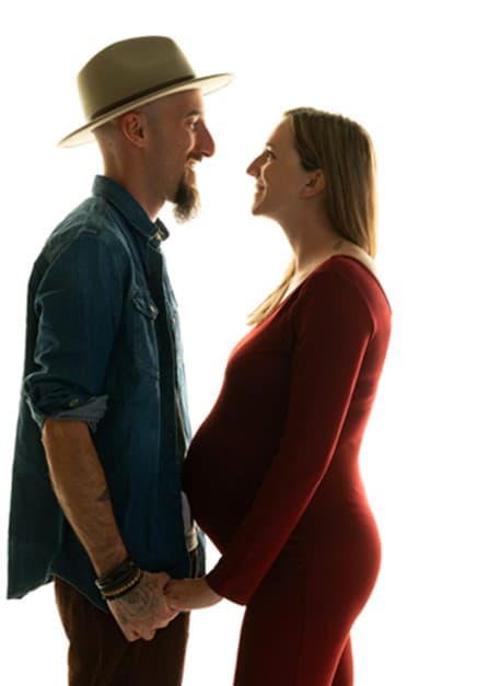 Future maman en robe rouge face à son conjoint en chemise en jean et chapeau, fond blanc