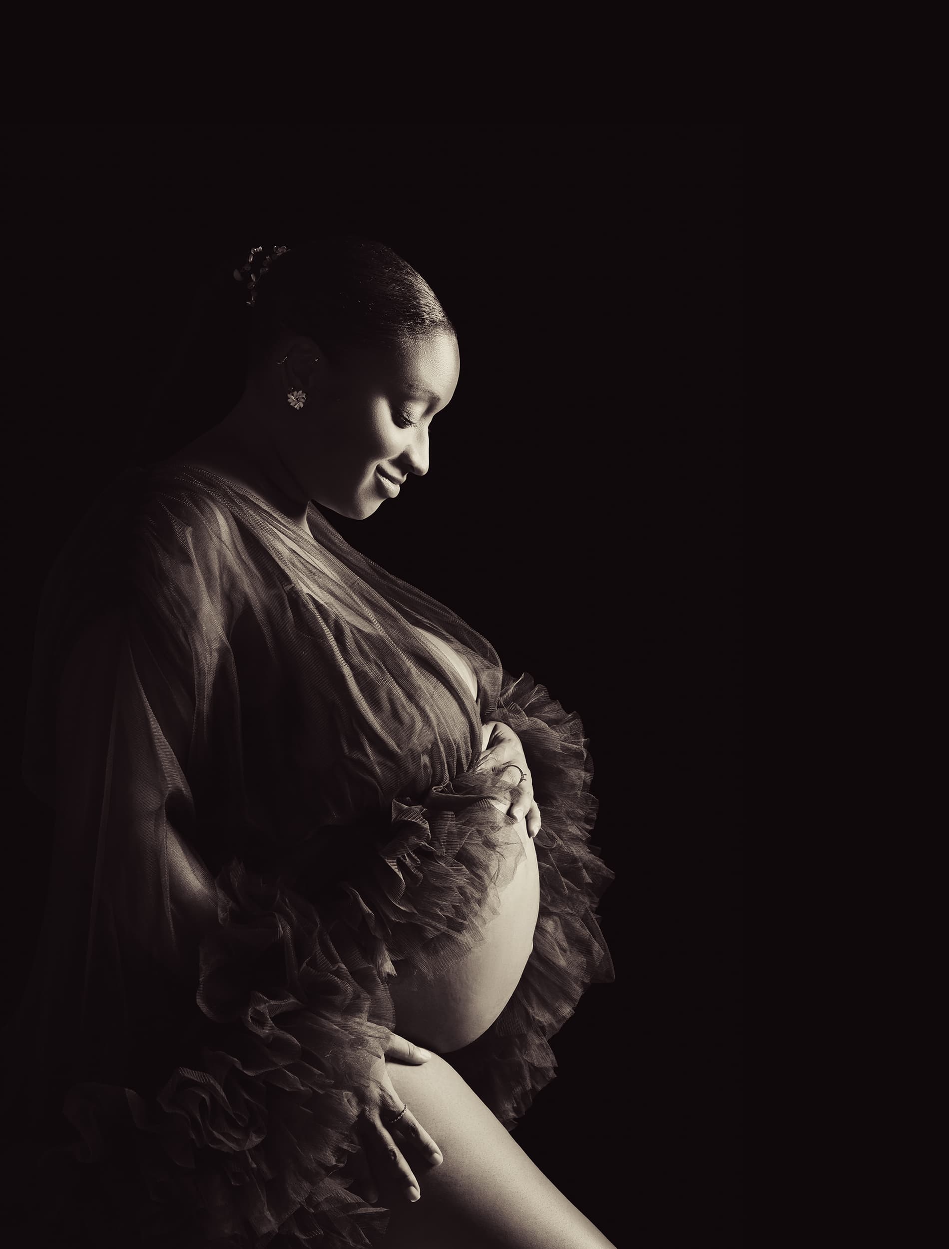 Future maman souriante, les yeux baissés vers son ventre, dans un voile de tulle sur fond noir