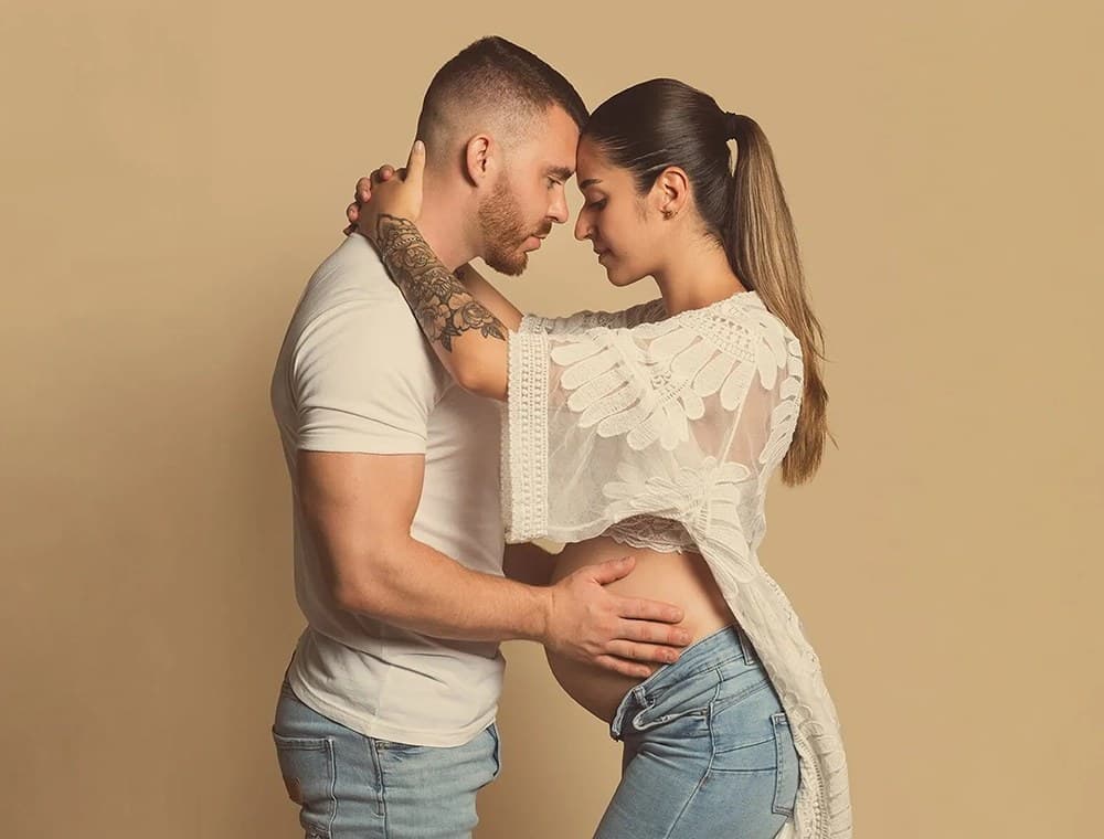 Futurs parents front contre front au studio, la main du papa sur le ventre, fond beige