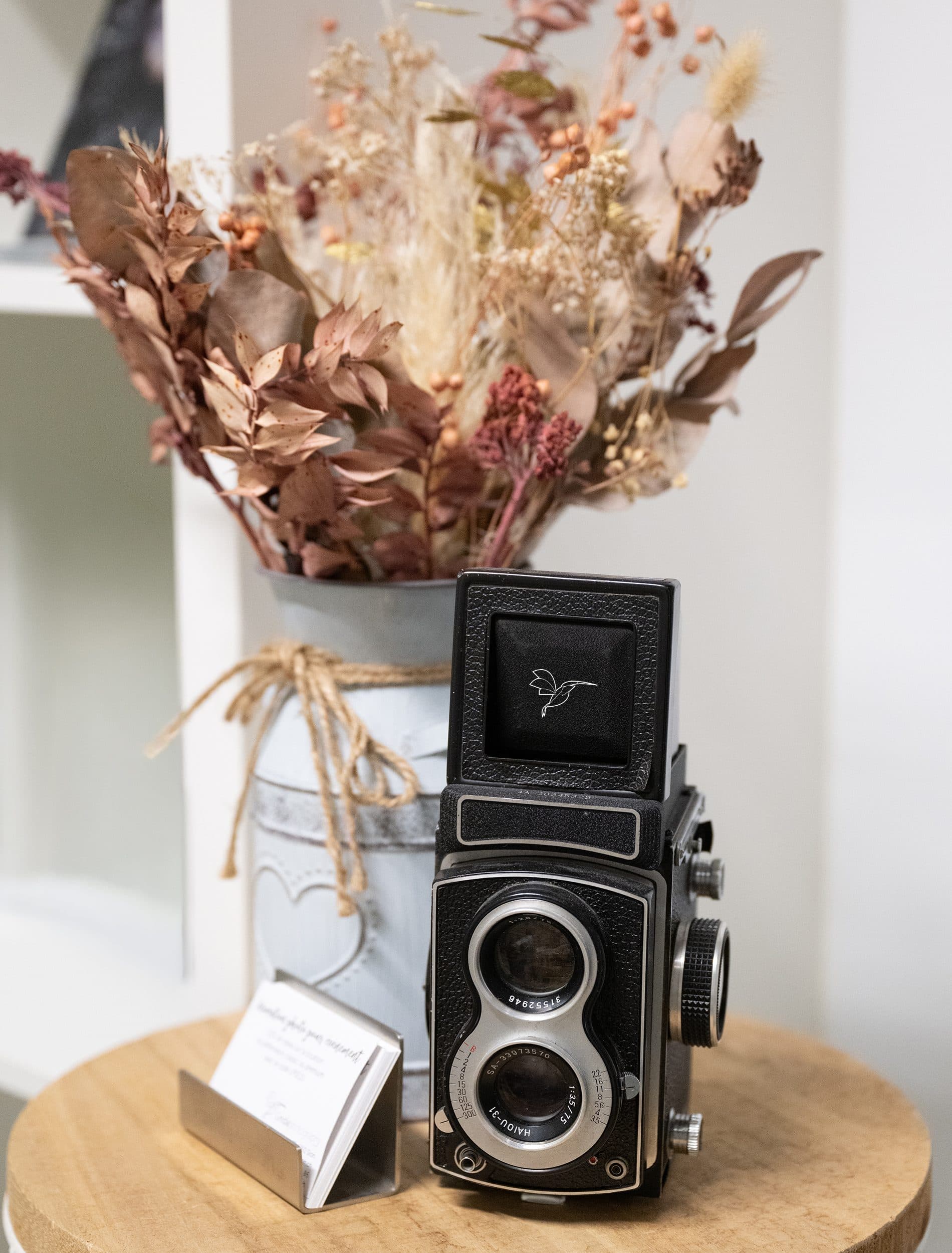 Un appareil photo ancien à deux objectifs et des cartes de visite, devant un bouquet de fleurs séchées, sur un tabouret en bois