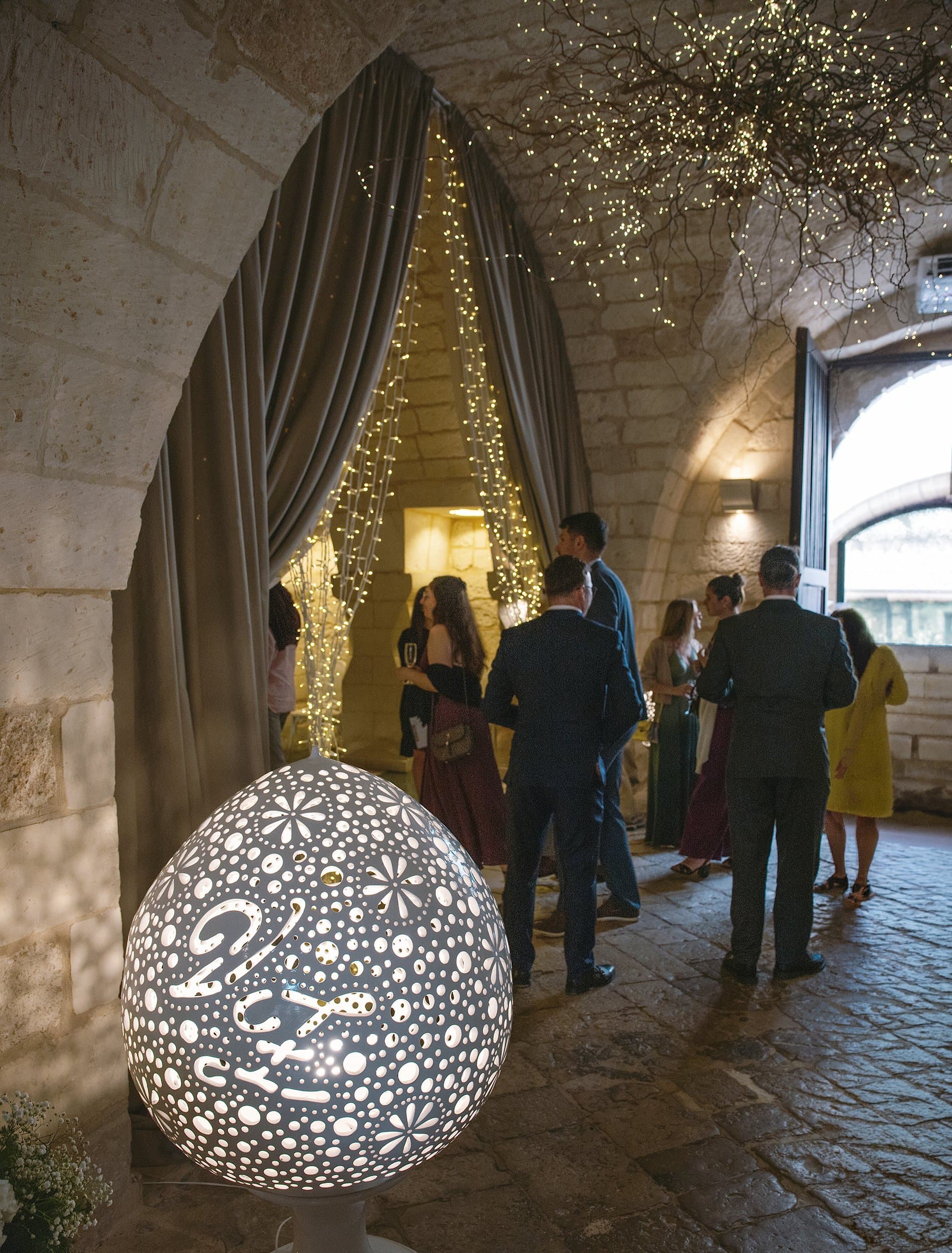 Apéritif sous des voûtes en pierre : des invités discutent près d’une lampe en forme d’œuf et d’un rideau de guirlandes lumineuses