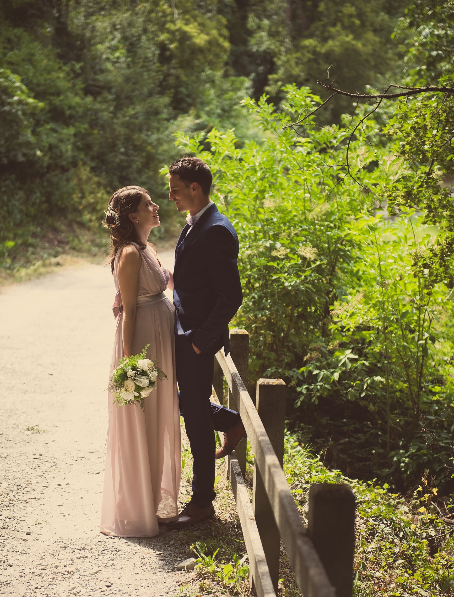 Les mariés face à face sur un chemin de forêt, la mariée en robe rose poudré, un bouquet blanc à la main