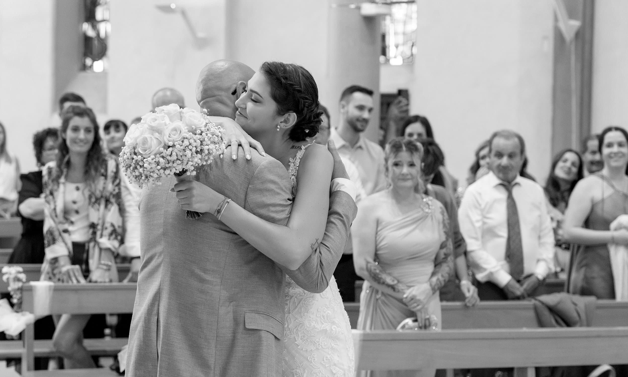 La mariée serre un proche dans ses bras dans l’allée de l’église, devant les invités debout, en noir et blanc