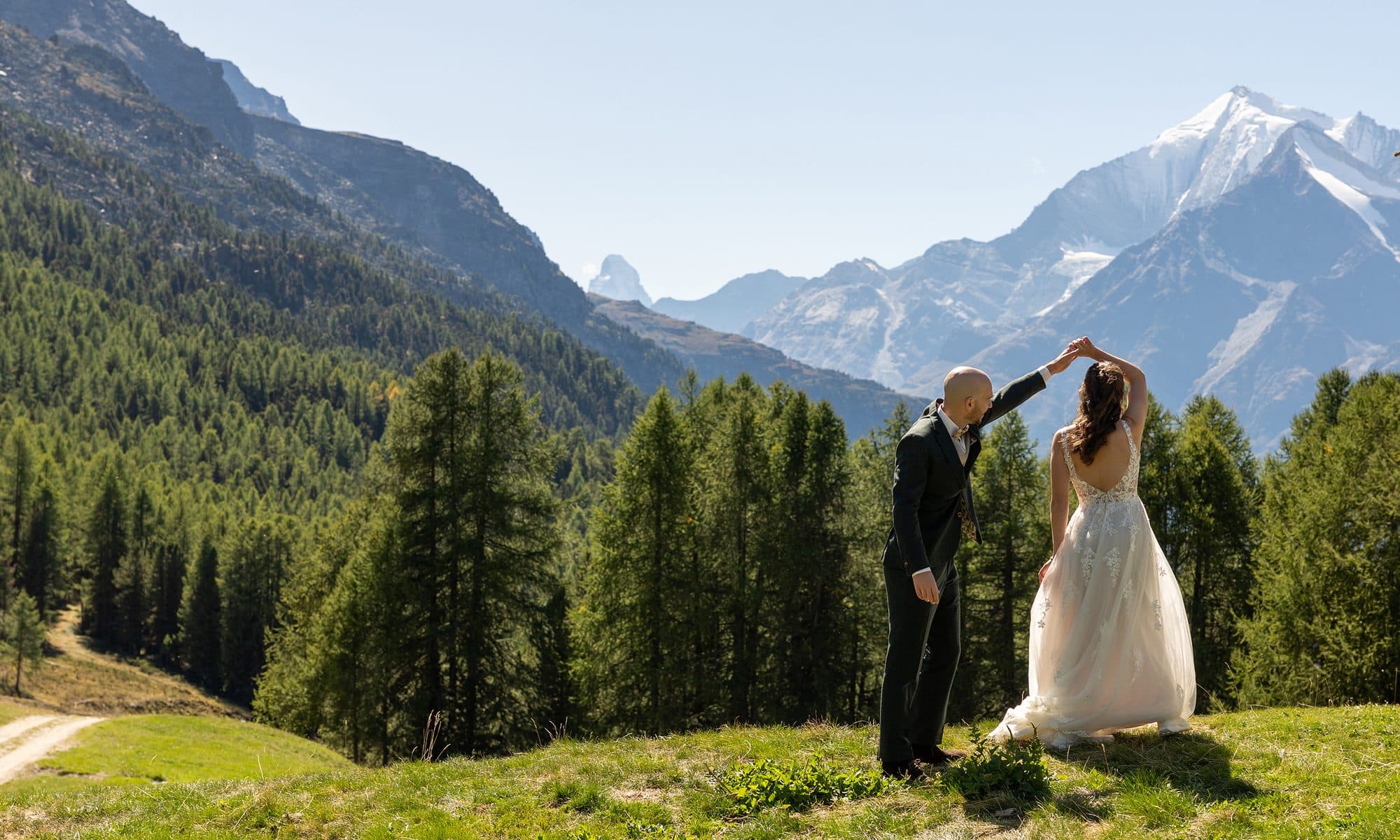 Les mariés dansent dans un pré de montagne, entre les mélèzes, face aux sommets enneigés