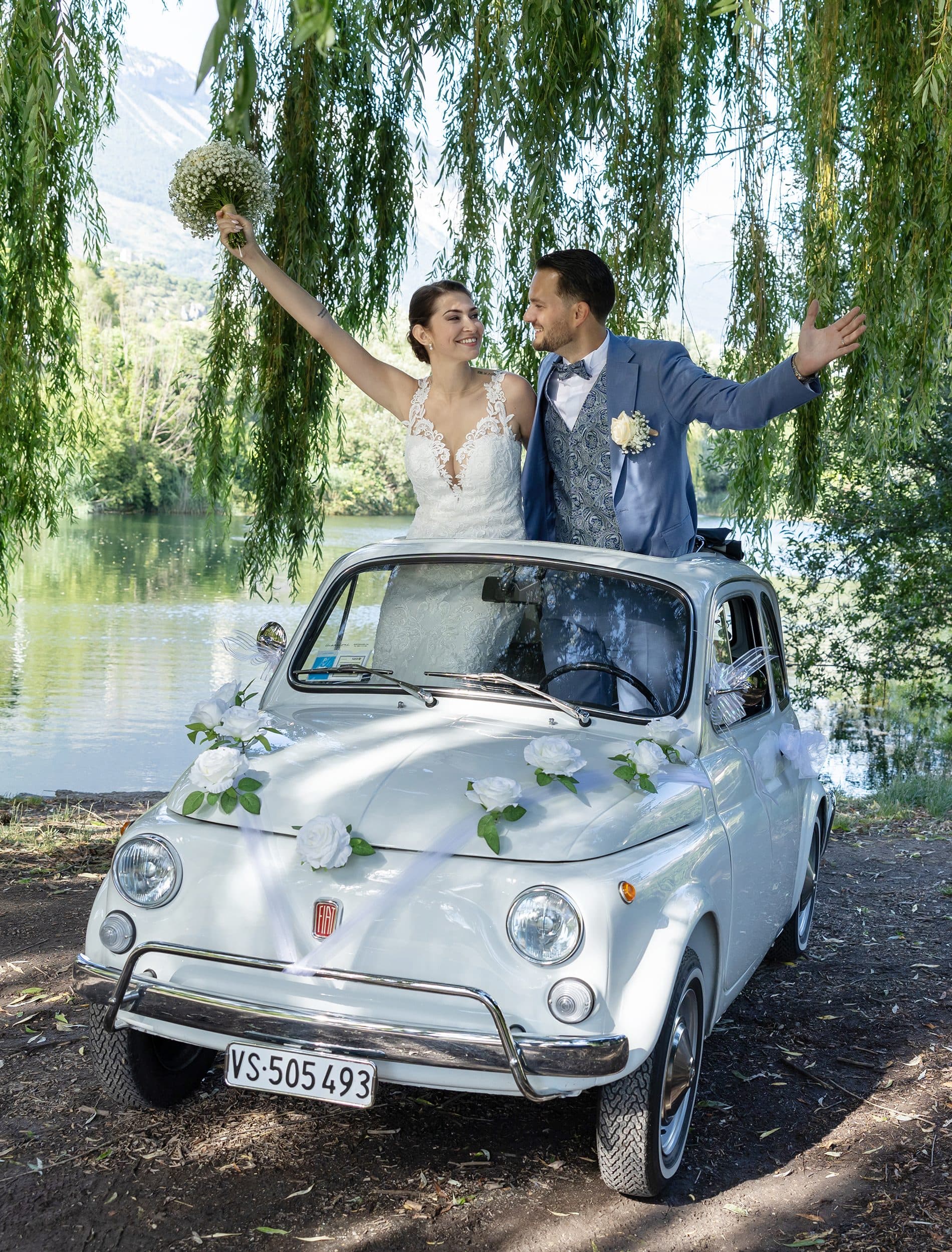 Les mariés lèvent les bras, debout dans une petite voiture blanche ancienne décorée de fleurs, sous un saule