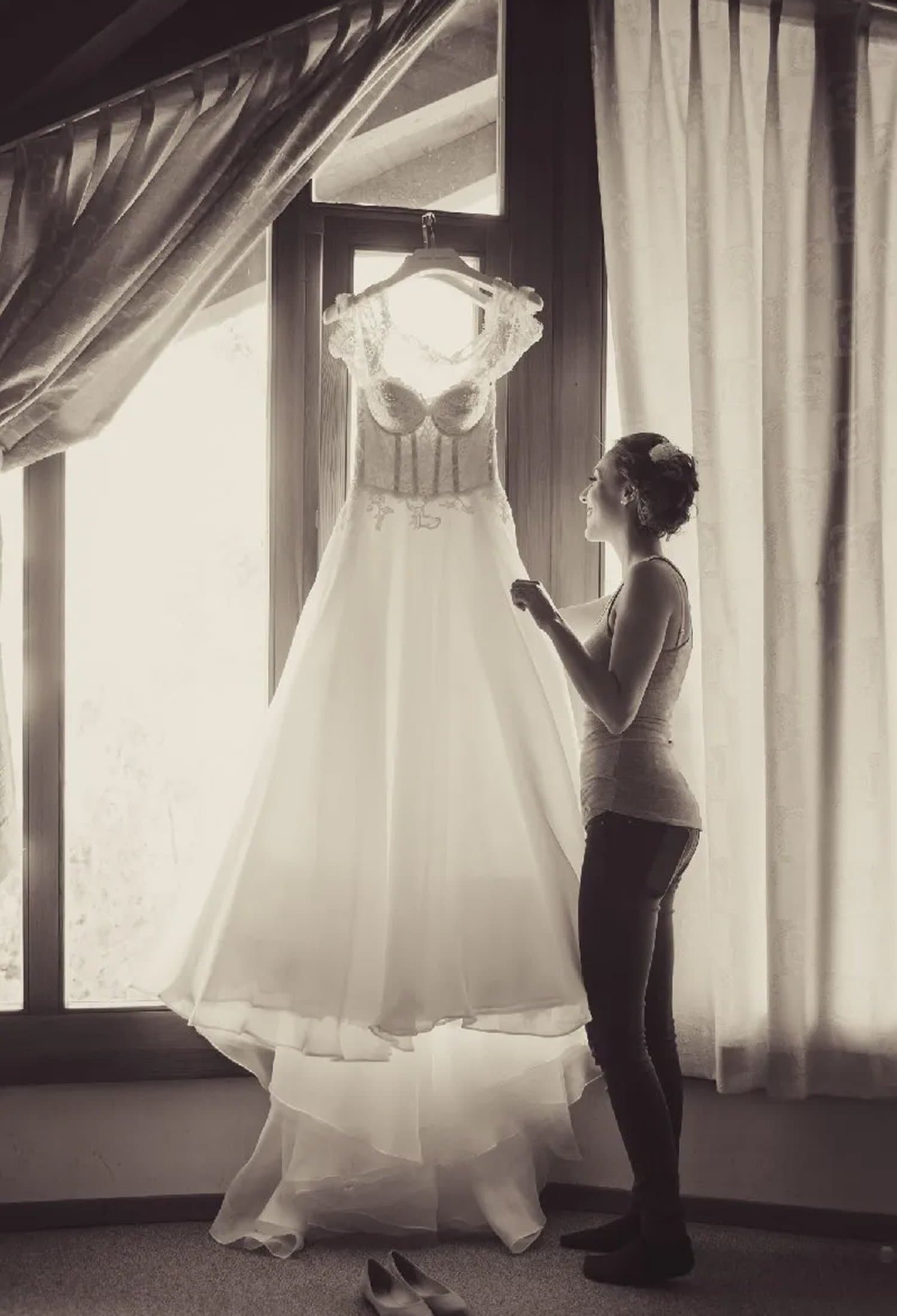 La mariée regarde sa robe suspendue devant une fenêtre, en noir et blanc