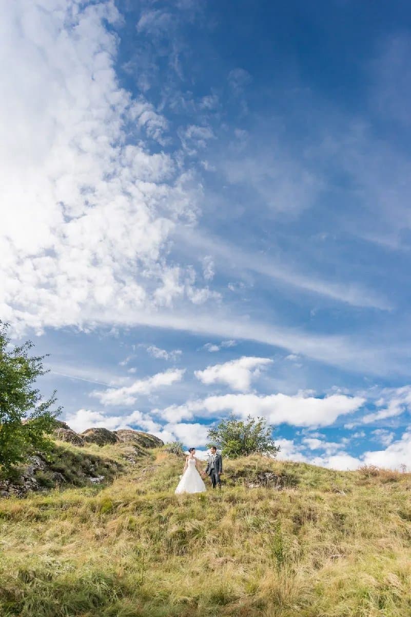 Les mariés, tout petits, sur une colline herbeuse sous un grand ciel bleu et nuageux