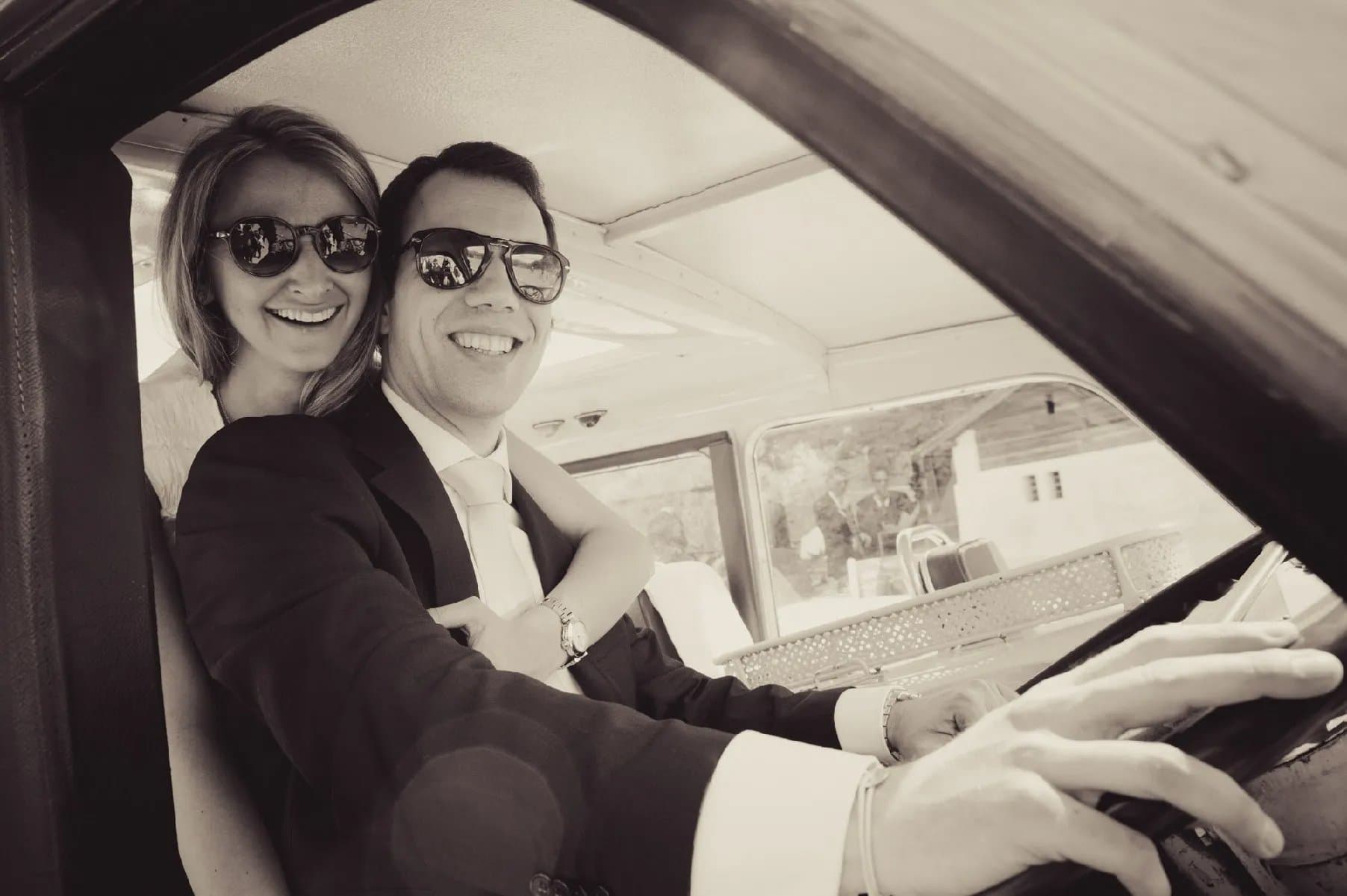 Les mariés en lunettes de soleil, souriants au volant d’une voiture ancienne, en tons sépia