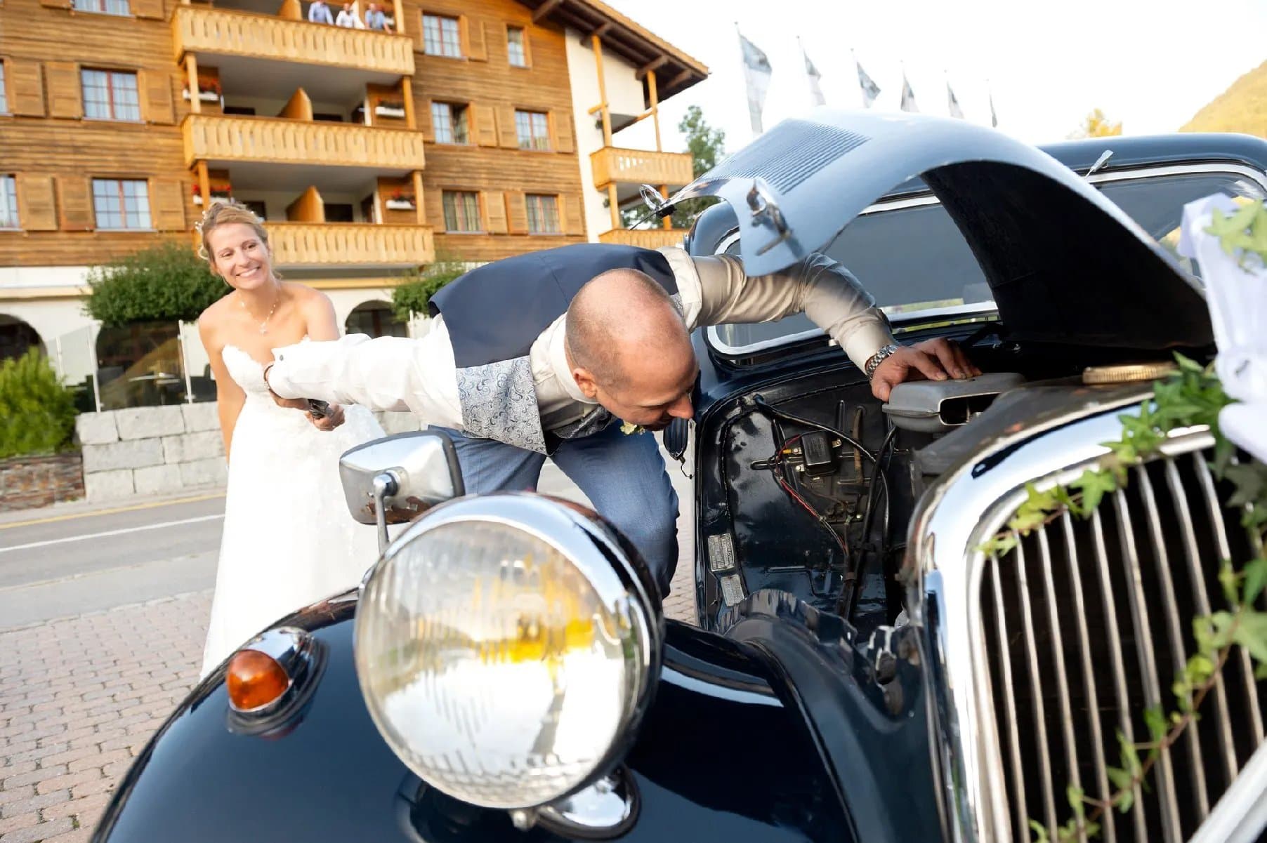 Le marié penché sous le capot d’une voiture ancienne, la mariée rit à côté de lui