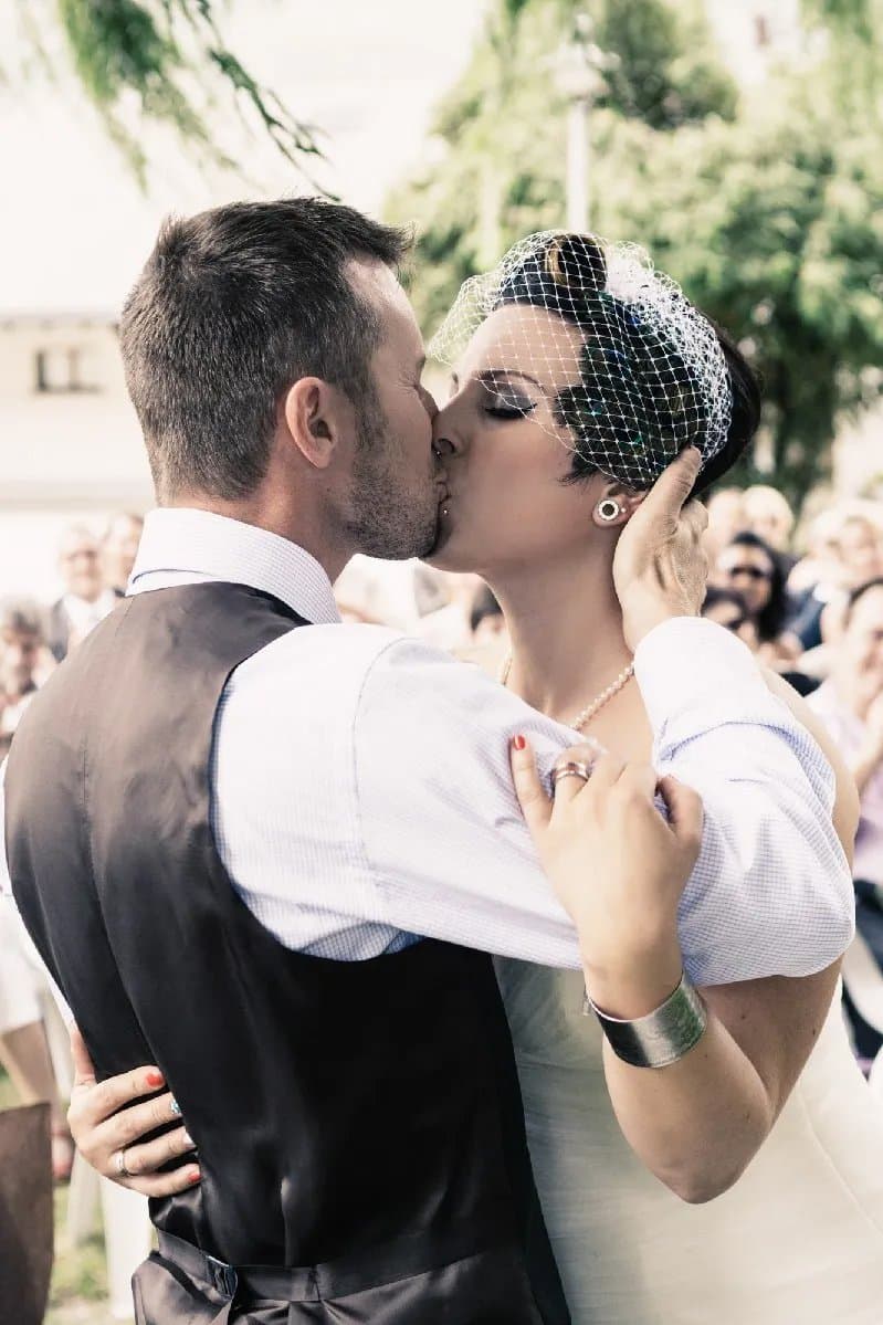 Baiser des mariés, la mariée coiffée d’une voilette, devant les invités
