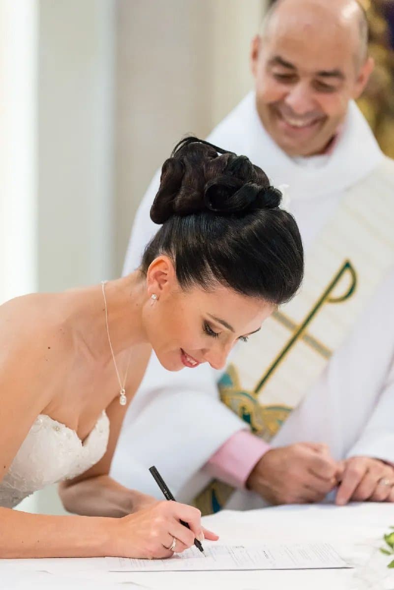 La mariée signe le registre, le prêtre sourit derrière elle