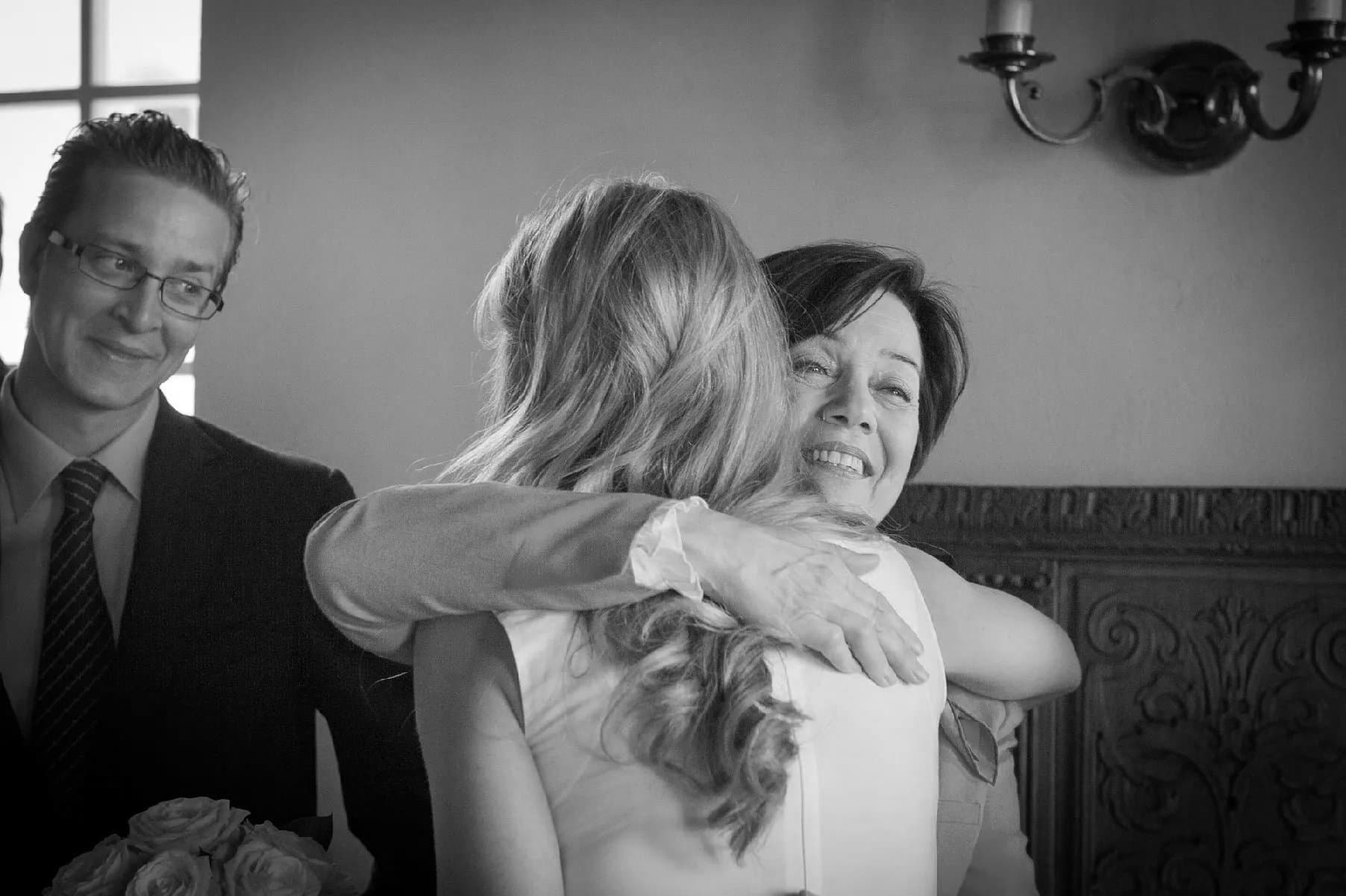 La mariée serrée dans les bras d’une invitée, un homme sourit à côté, en noir et blanc