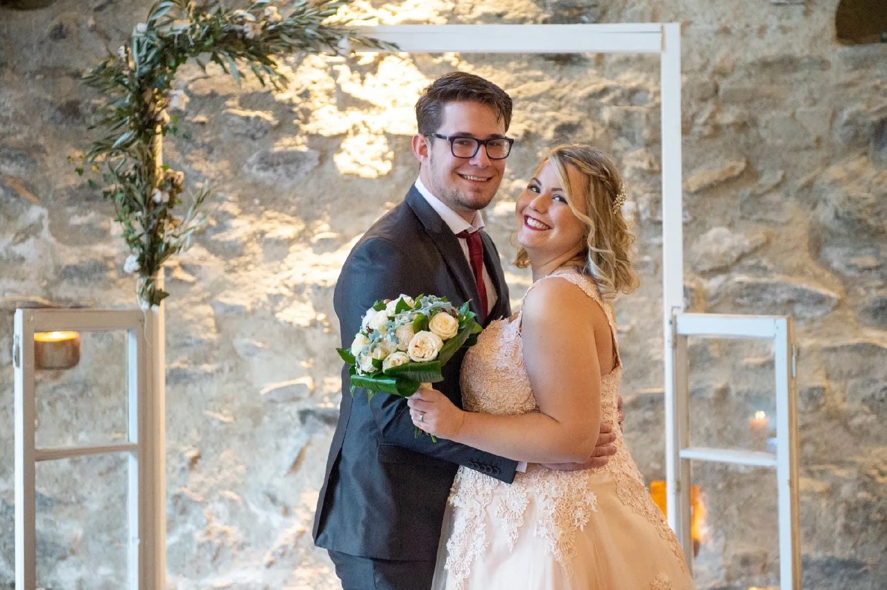 Les mariés enlacés devant une arche blanche ornée de verdure et un mur en pierre