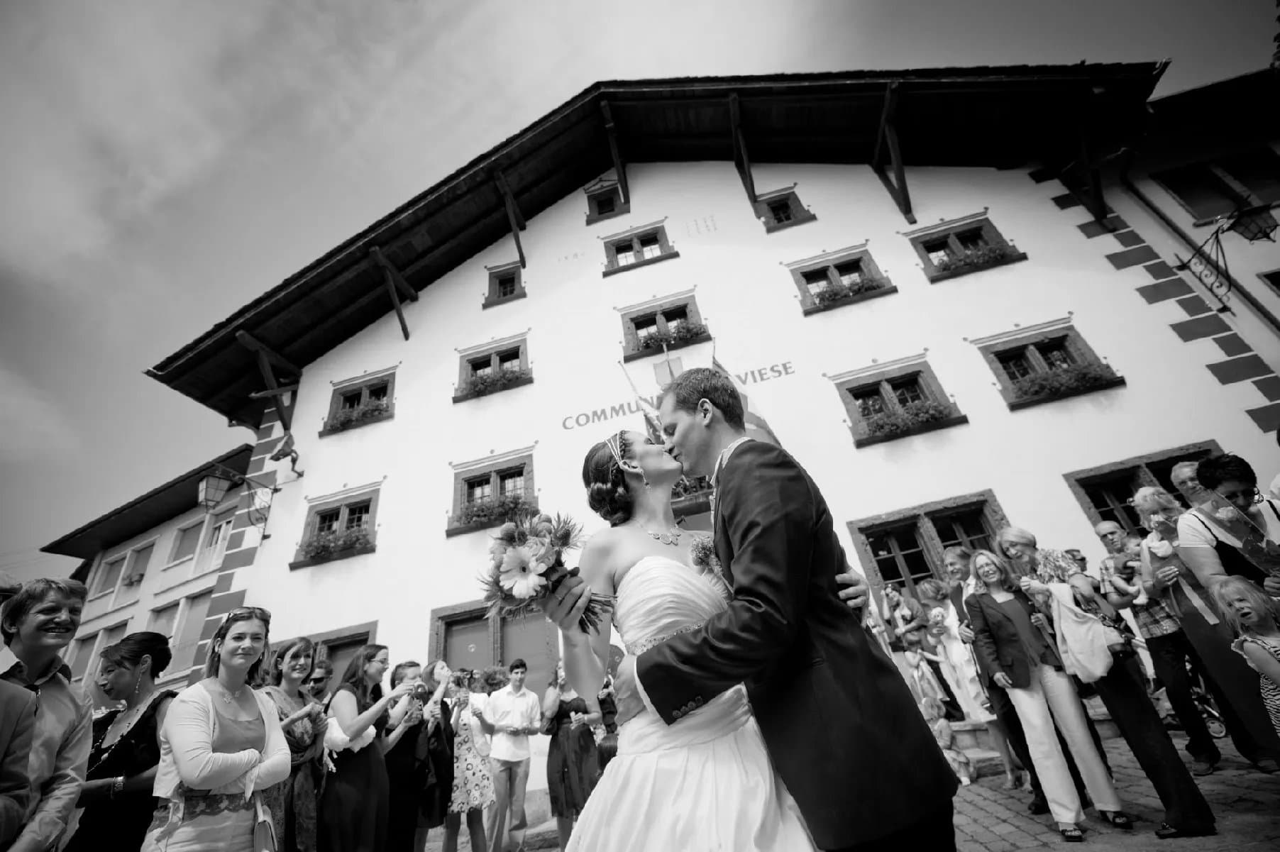 Baiser des mariés devant la maison de commune de Savièse, entourés de leurs invités, en noir et blanc