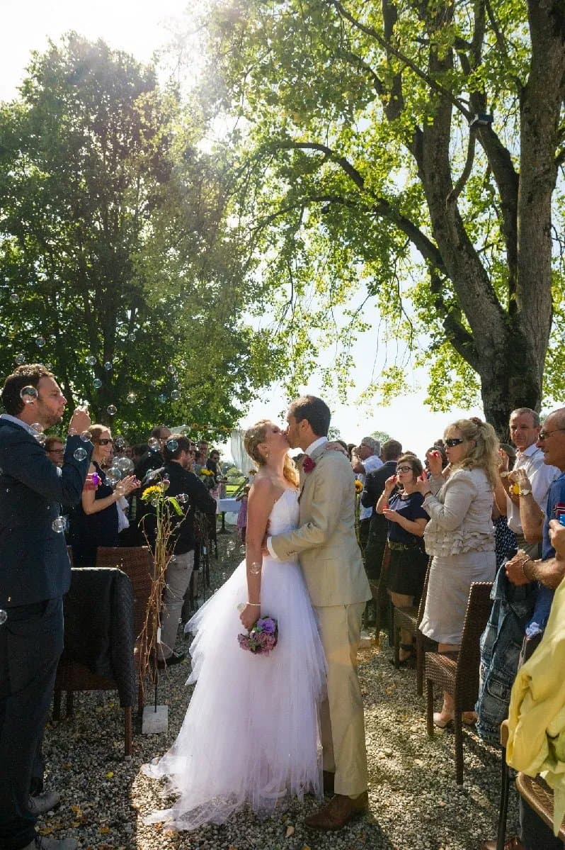 Baiser des mariés au bout de l’allée d’une cérémonie en plein air, sous les arbres