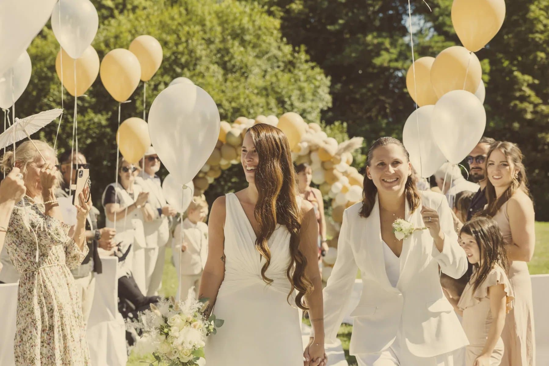 Deux mariées main dans la main au milieu de leurs invités et de ballons dorés et blancs
