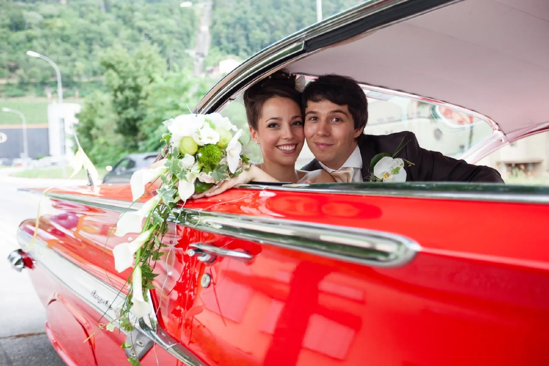Les mariés à la fenêtre d’une voiture américaine rouge décorée de fleurs blanches