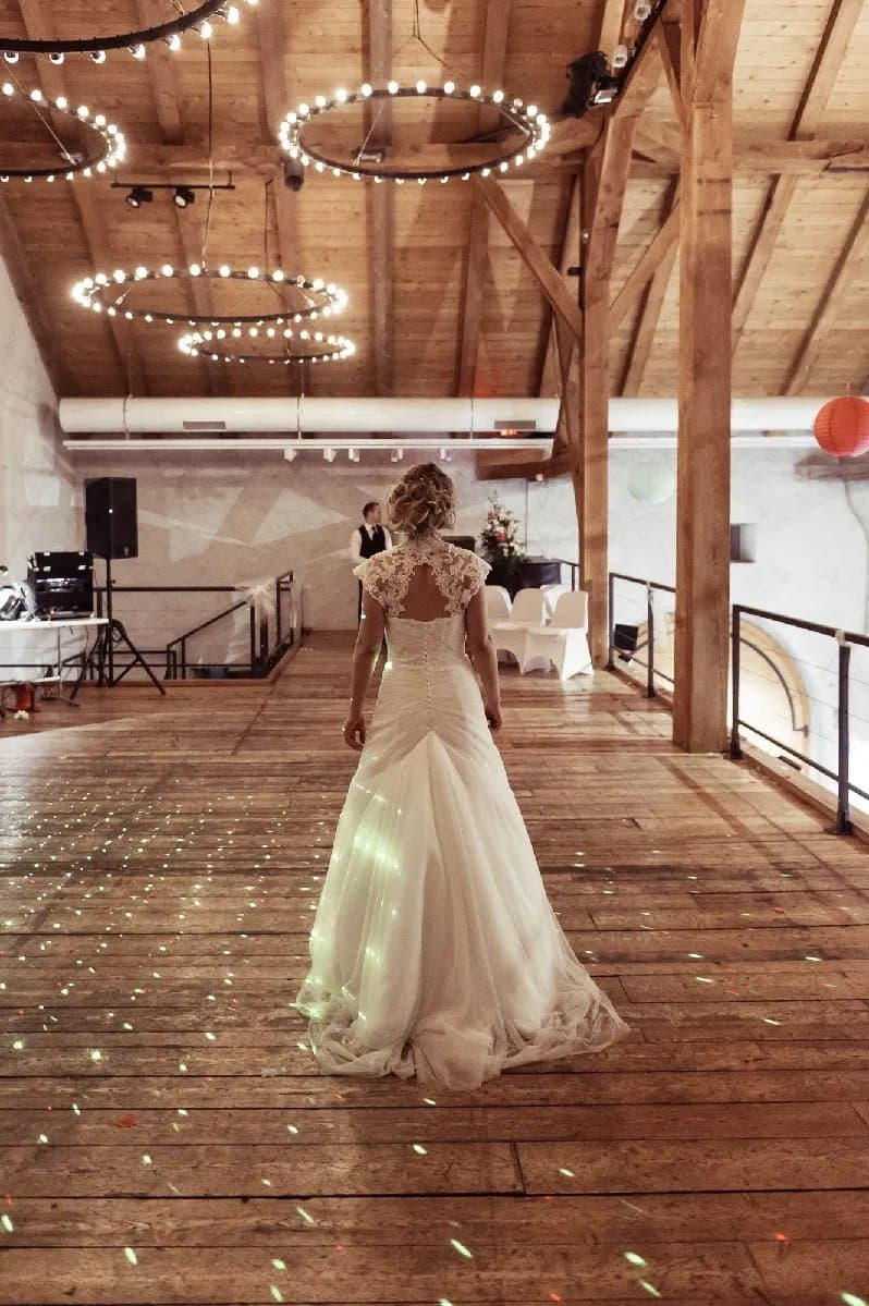 La mariée, de dos, traverse une salle à la charpente en bois éclairée de cercles lumineux