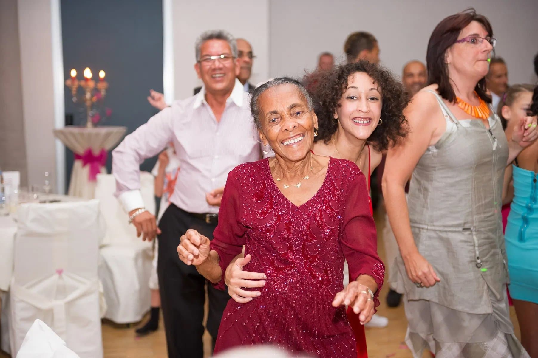 Les invités dansent, une aînée en robe bordeaux sourit au premier plan