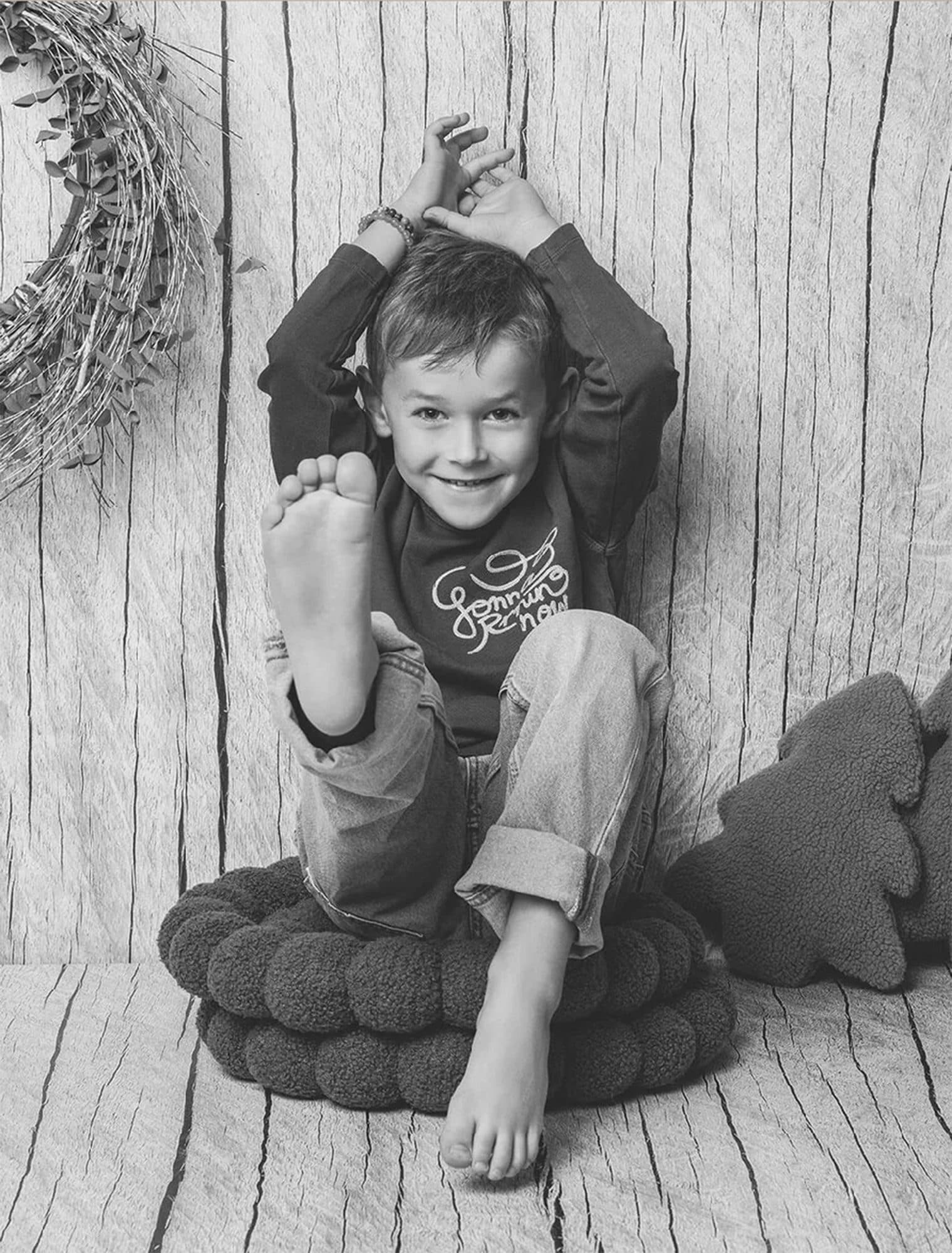 Décor Bois Clair et Vert Olive : un petit garçon rieur assis sur un coussin tressé, un pied levé, en noir et blanc