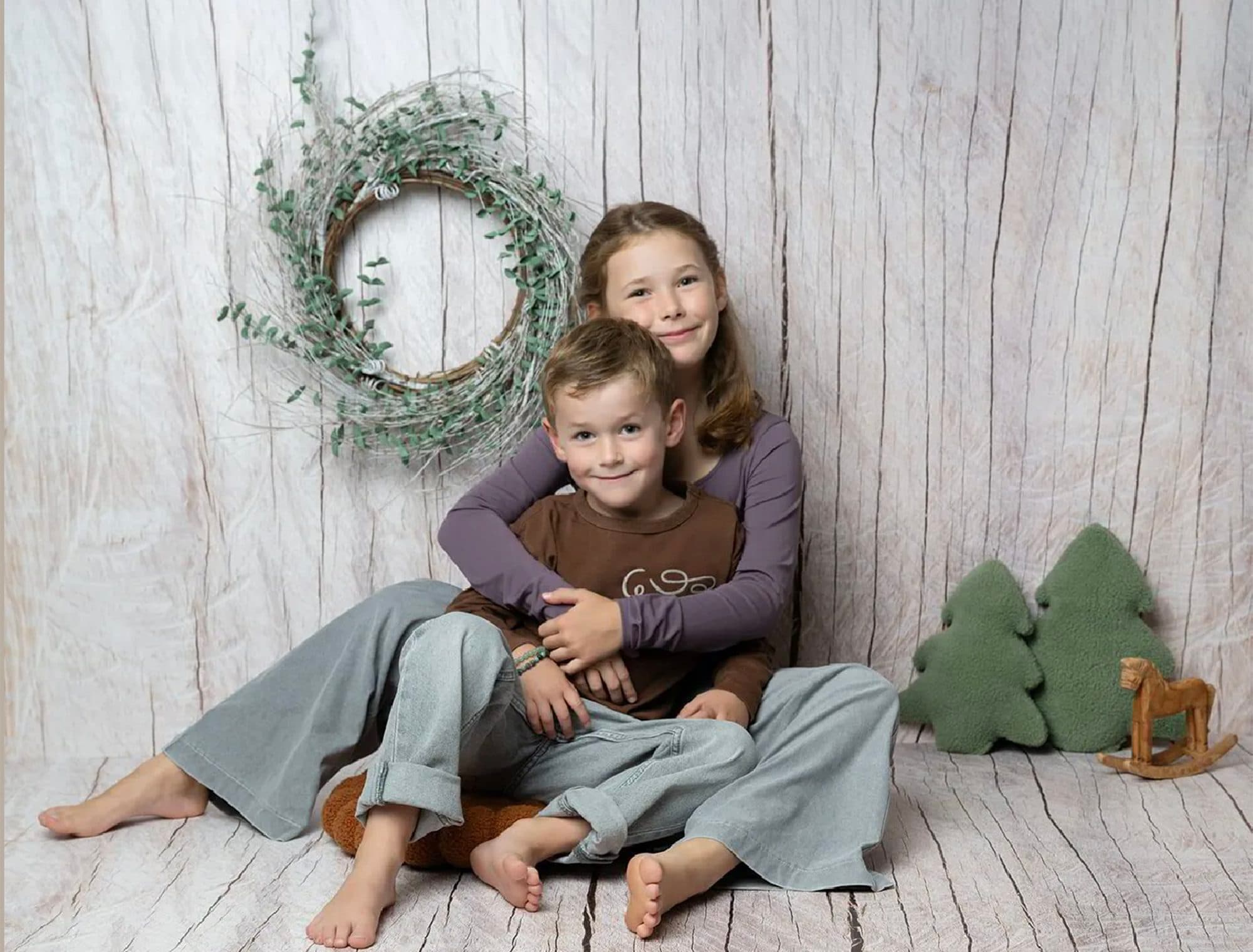 Décor Bois Clair et Vert Olive : une grande sœur enlace son petit frère, assis devant un mur en bois blanchi et une couronne de verdure