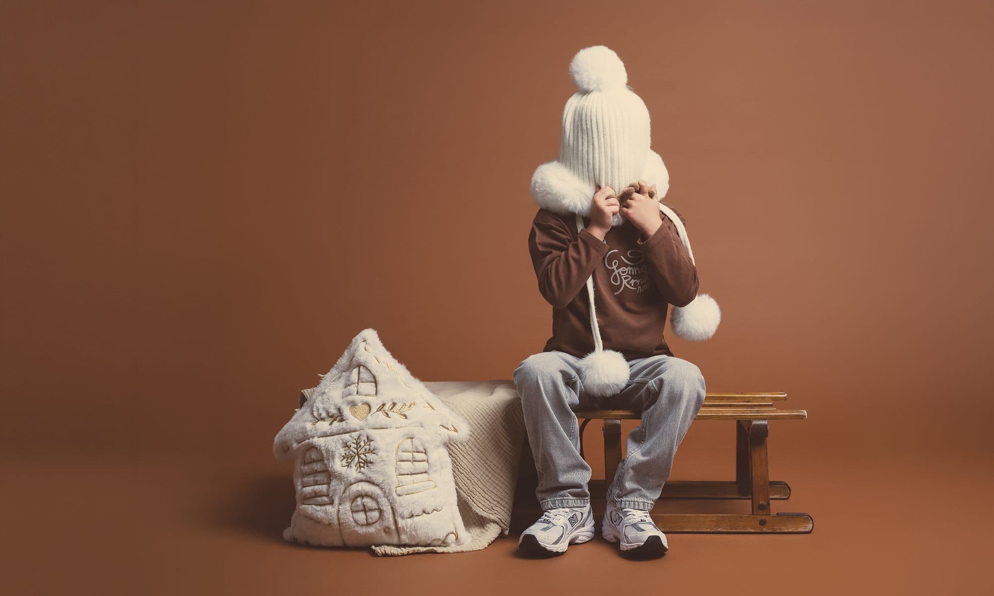Un petit garçon en bonnet blanc à pompon assis sur une luge en bois, à côté d’une maison en tissu, devant un fond brun cacao